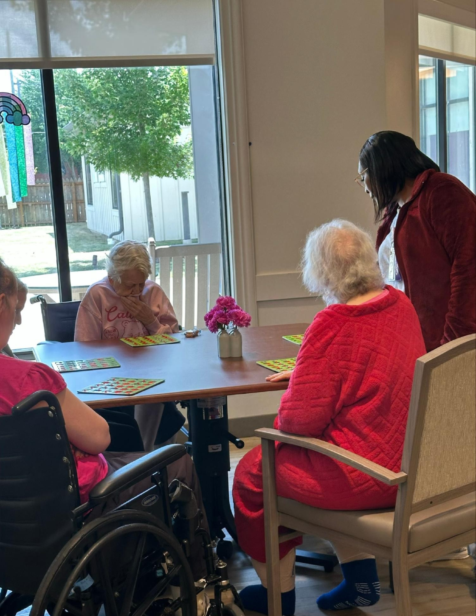 A group of elderly women sitting around a dining table playing bingo, with a caregiver or staff member standing nearby. The room is well-lit with natural sunlight coming through large windows.