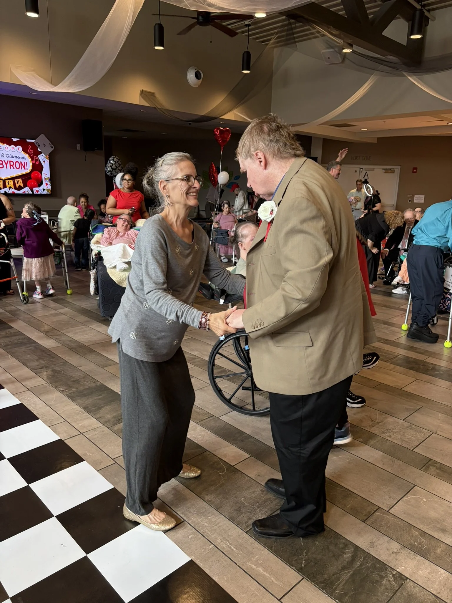 An elderly woman and an elderly man shake hands and smile at each other in a decorated indoor event space, with several people in the background, some using wheelchairs, and a large screen displaying the name Byron.