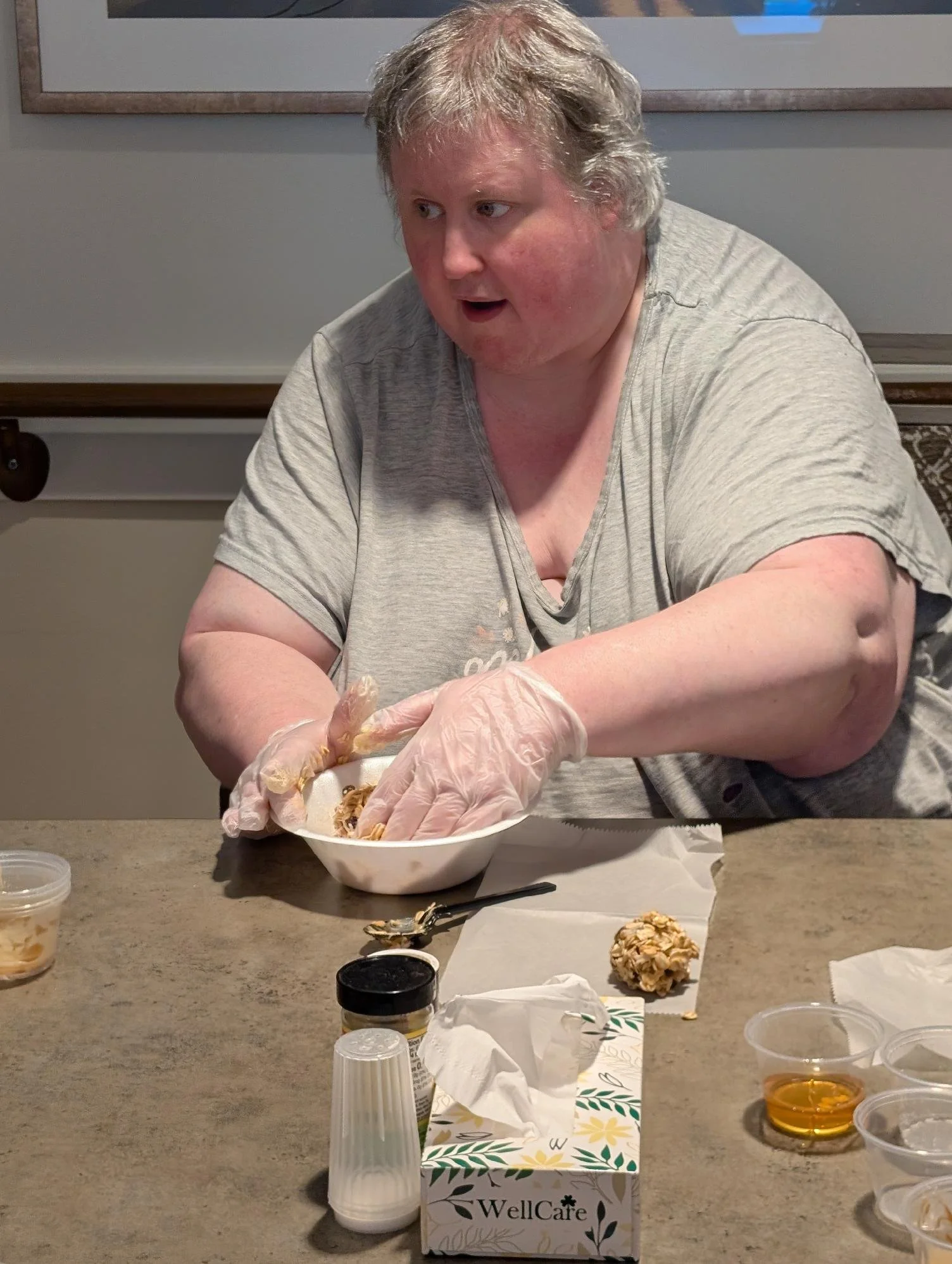 Person with gray hair wearing gloves, sitting at a table and handling food in a bowl, surrounded by food containers, cups, and a tissue box labeled WellCare.