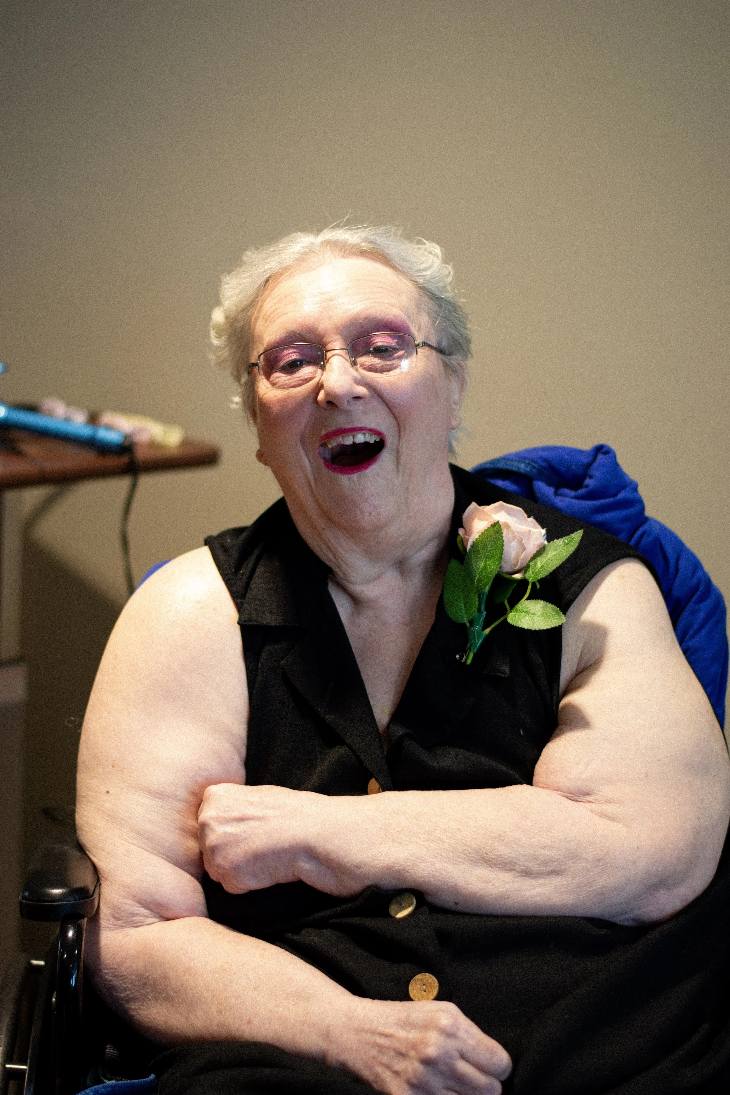 An elderly woman with gray hair and glasses is smiling with her mouth open. She is sitting in a wheelchair, wearing a black sleeveless top, and has a pink rose with green leaves pinned to her clothing. Her arms are crossed in front of her, and she ap