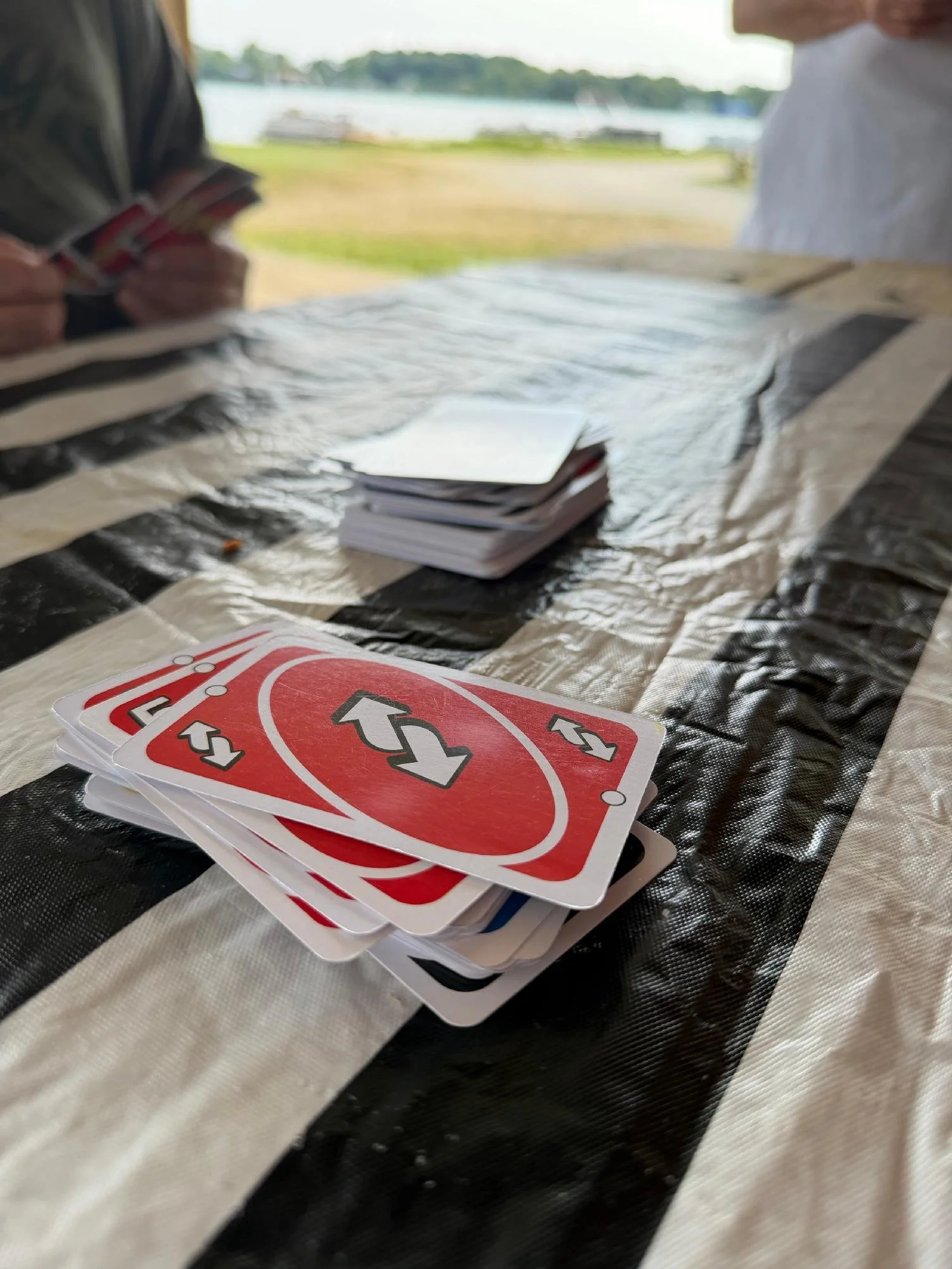 Close-up of a deck of Uno playing cards on a striped black and beige plastic tablecloth, with a blurred outdoor background featuring water and trees.
