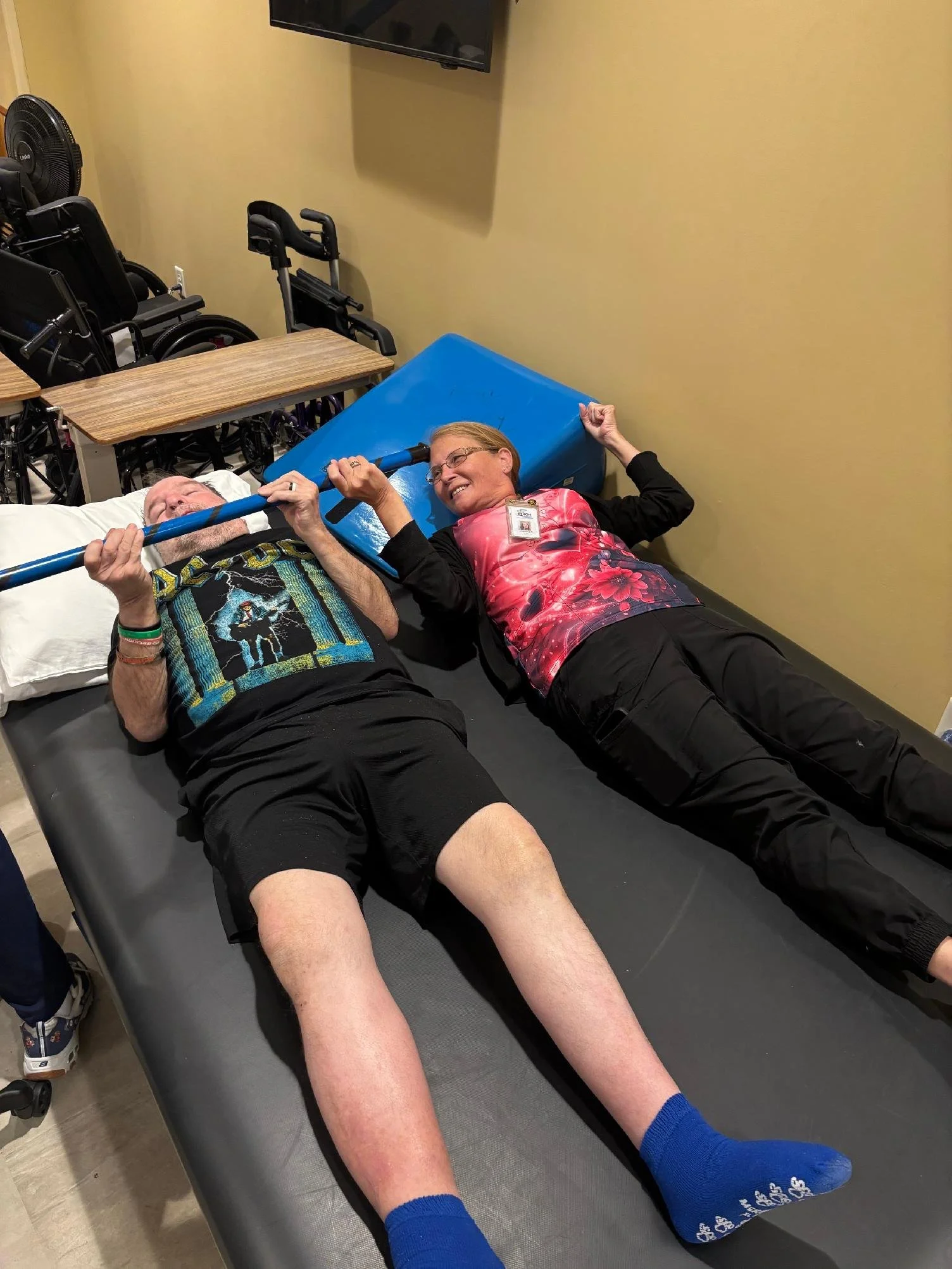 An elderly man and a woman lying on a medical examination table, holding a pole above their heads as they smile. The room has a beige wall, a mounted TV, and wheelchair equipment in the background.