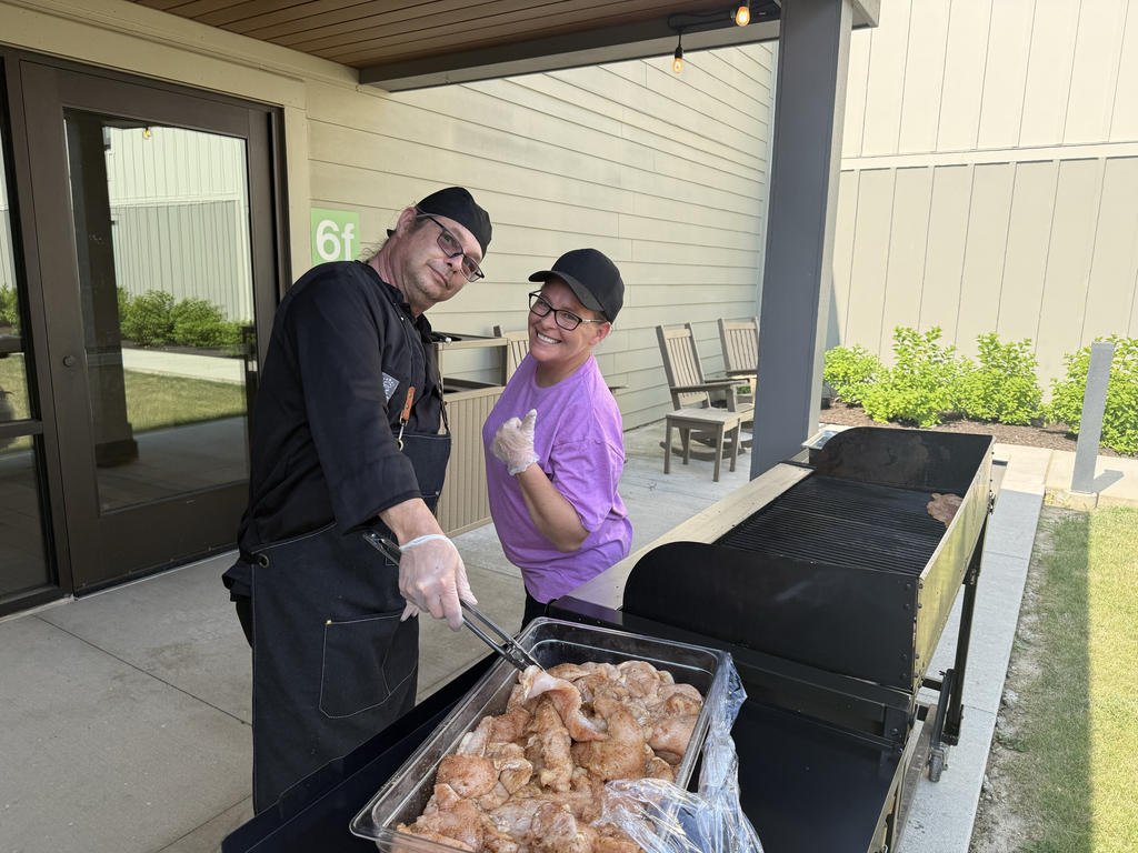 Two people dressed in casual clothing and aprons, standing next to a grill outdoors, preparing chicken pieces. One person is holding tongs. They are smiling and posing for the photo. There are chairs, greenery, and a building in the background.