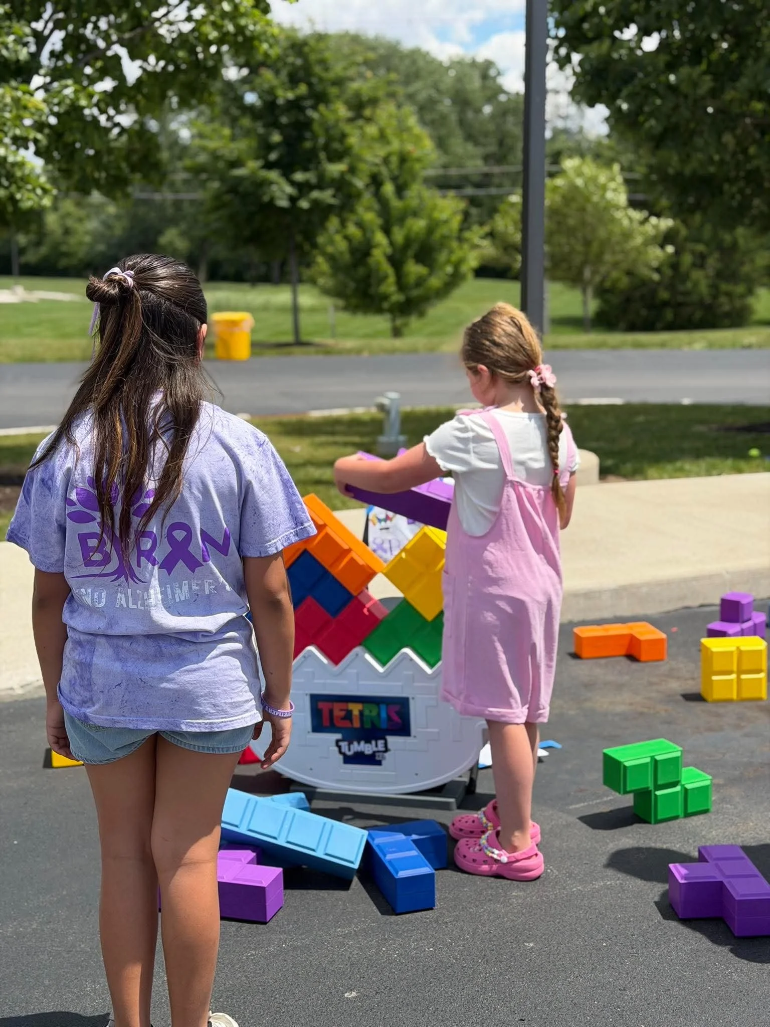Two young girls are playing with large colorful plastic Tetris blocks outdoors on a sunny day. One girl has long brown hair tied back with a pink scrunchie and is wearing a pink dress and pink Crocs. The other girl has long dark hair in a loose bun w