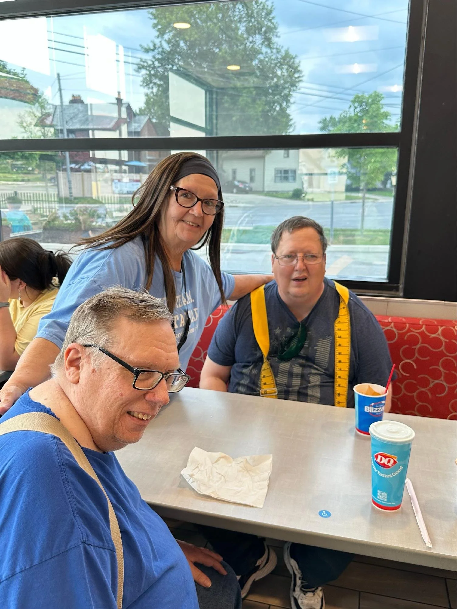 Four people sitting at a table inside a Dairy Queen restaurant. A woman in a blue shirt and glasses is smiling at the camera, while a man in a navy shirt with a yellow measuring tape around his neck looks surprised. The person in front of them has gray hair and glasses and is smiling. On the table are two Dairy Queen cups, a napkin, and a drink with a straw. Outside the window, trees, houses, and power lines are visible.