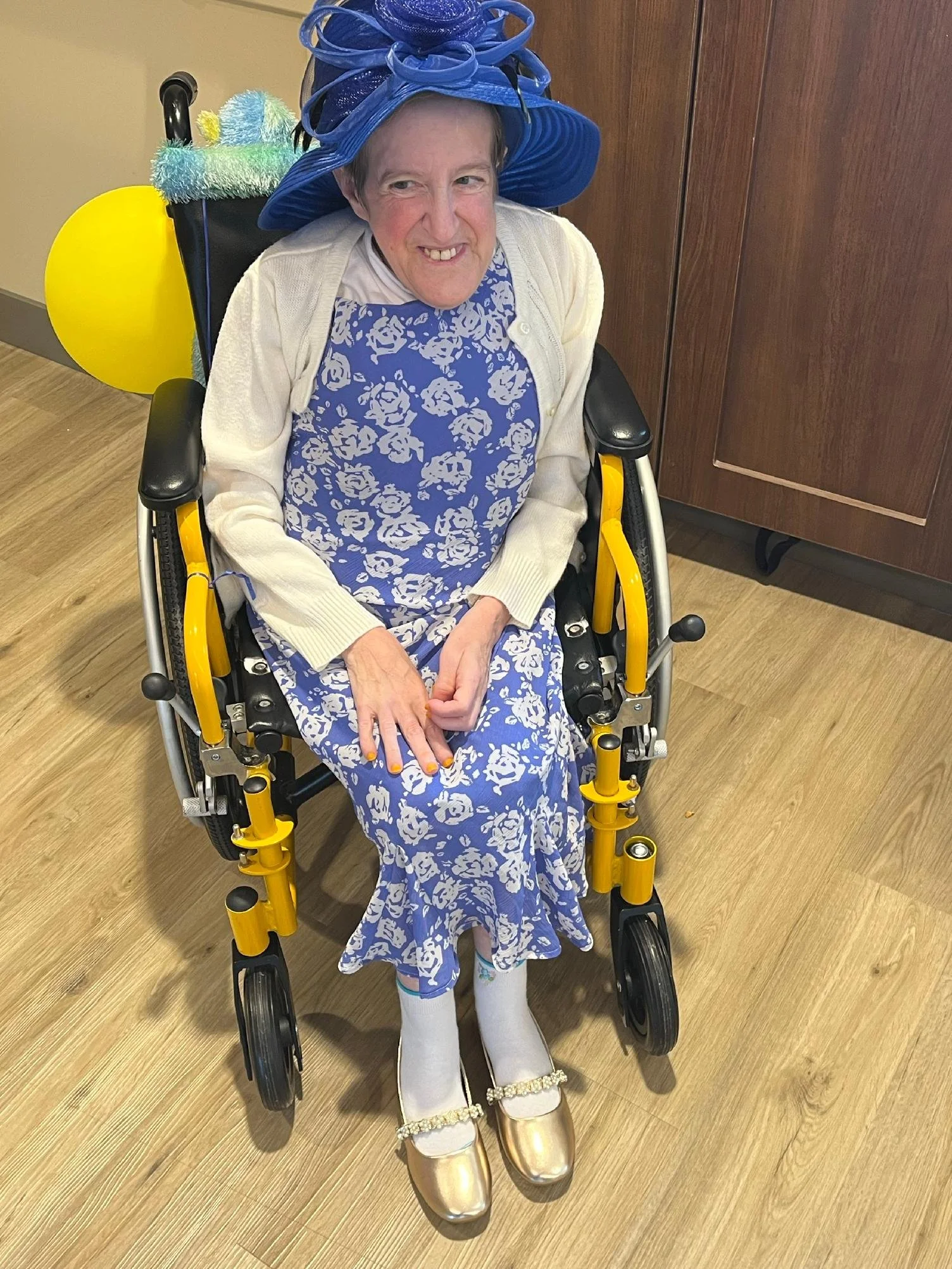 An elderly woman sitting in a yellow wheelchair wearing a blue floral dress, a cream cardigan, white tights, and shiny gold shoes. She is also wearing a large, elaborate blue hat and has her nails painted orange. She is smiling and appears to be at a