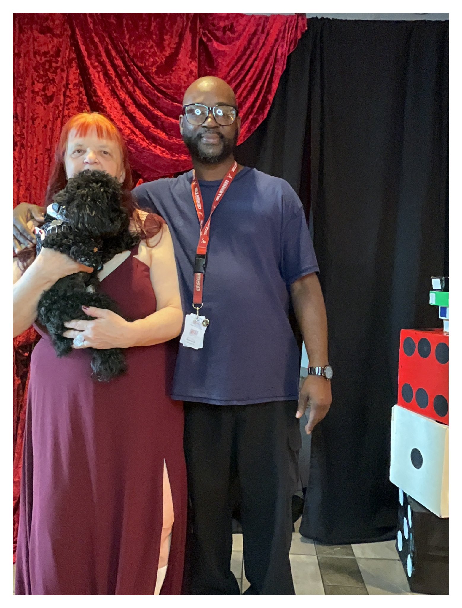 Two people standing side by side in front of a red and black curtain, with only the lower half of a woman and the upper half of a man visible. The woman has red hair and is holding a small black dog, while the man is wearing glasses and a navy shirt 