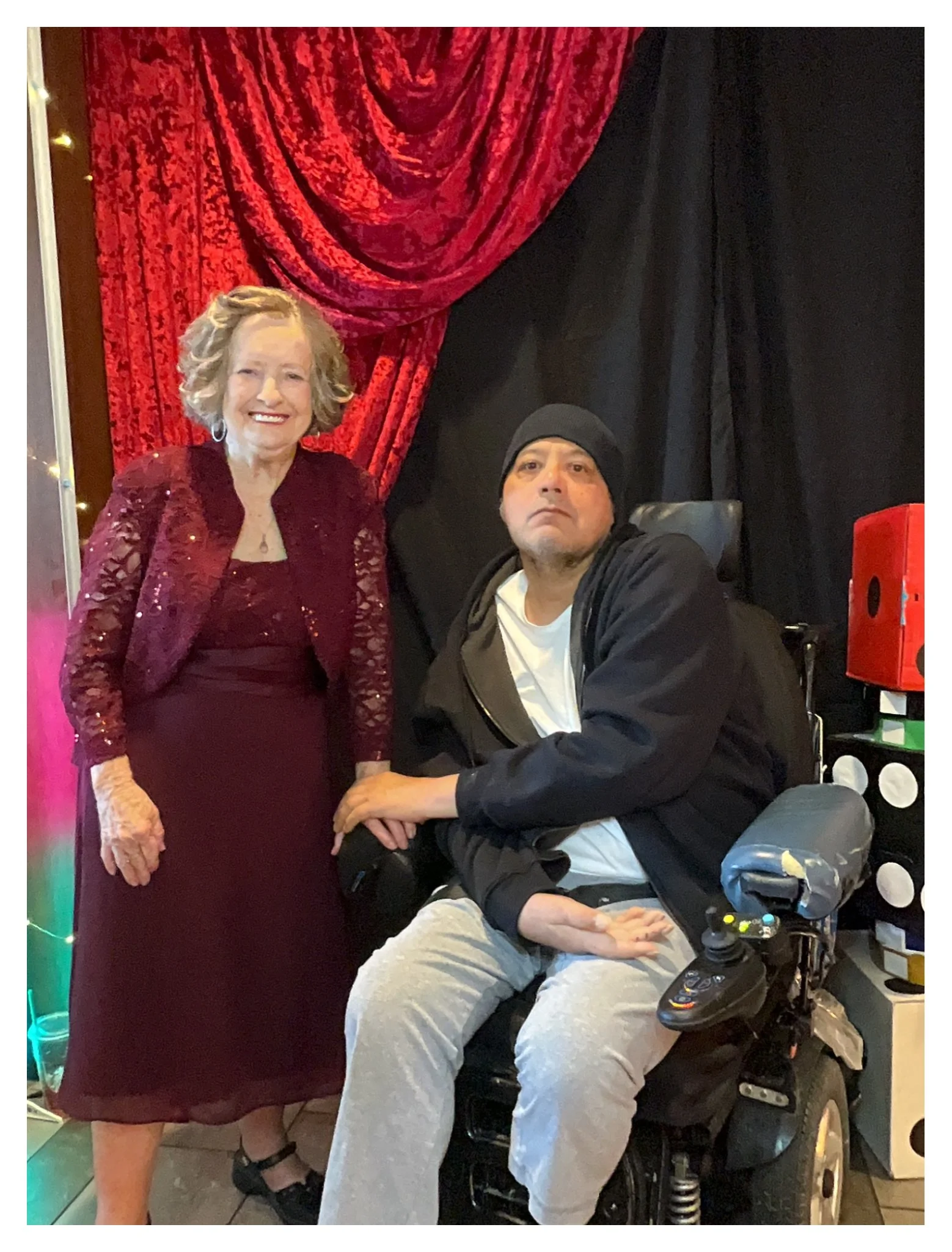A woman in a red dress and jacket standing next to a man in a wheelchair. The woman has light-colored, curly hair and is smiling, while the man is wearing a black beanie and looking serious. The background features red and black curtains.