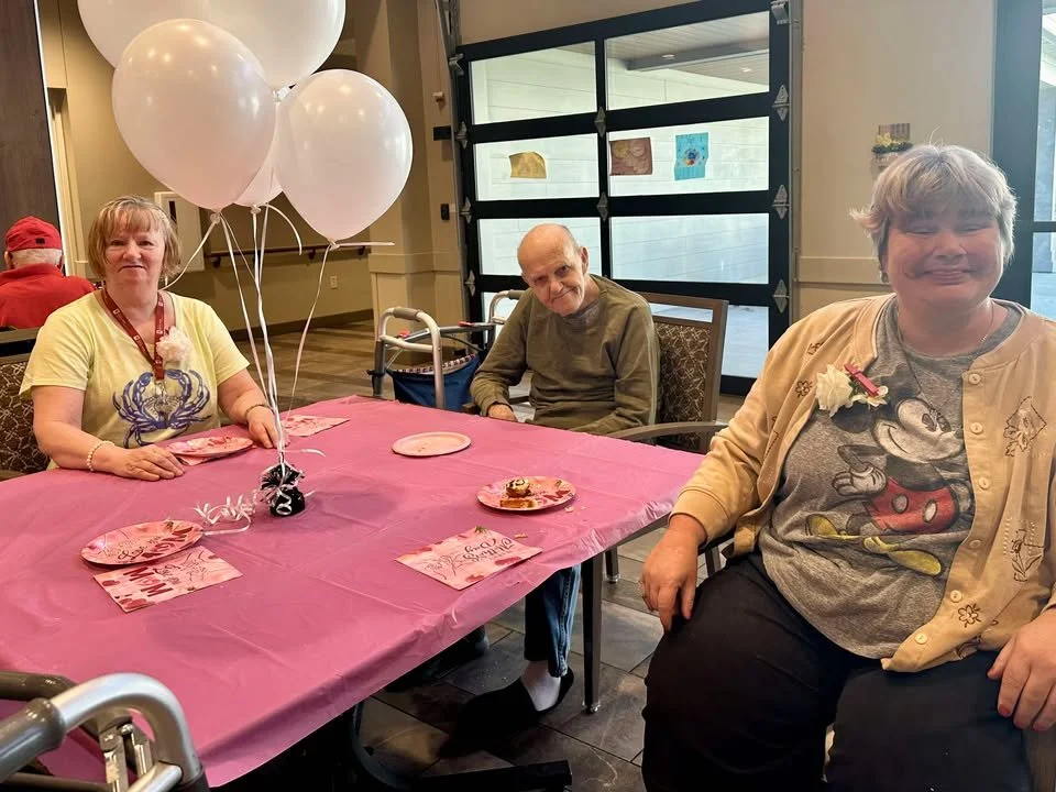 Two women and one elderly man sitting at a table decorated with pink tablecloth and balloons, celebrating a special occasion with cake and greeting cards.