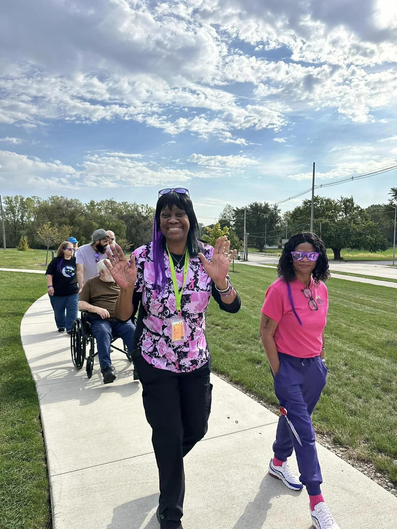 People walking outdoors on a sidewalk in a park, smiling and waving at the camera on a sunny day with a partly cloudy sky.