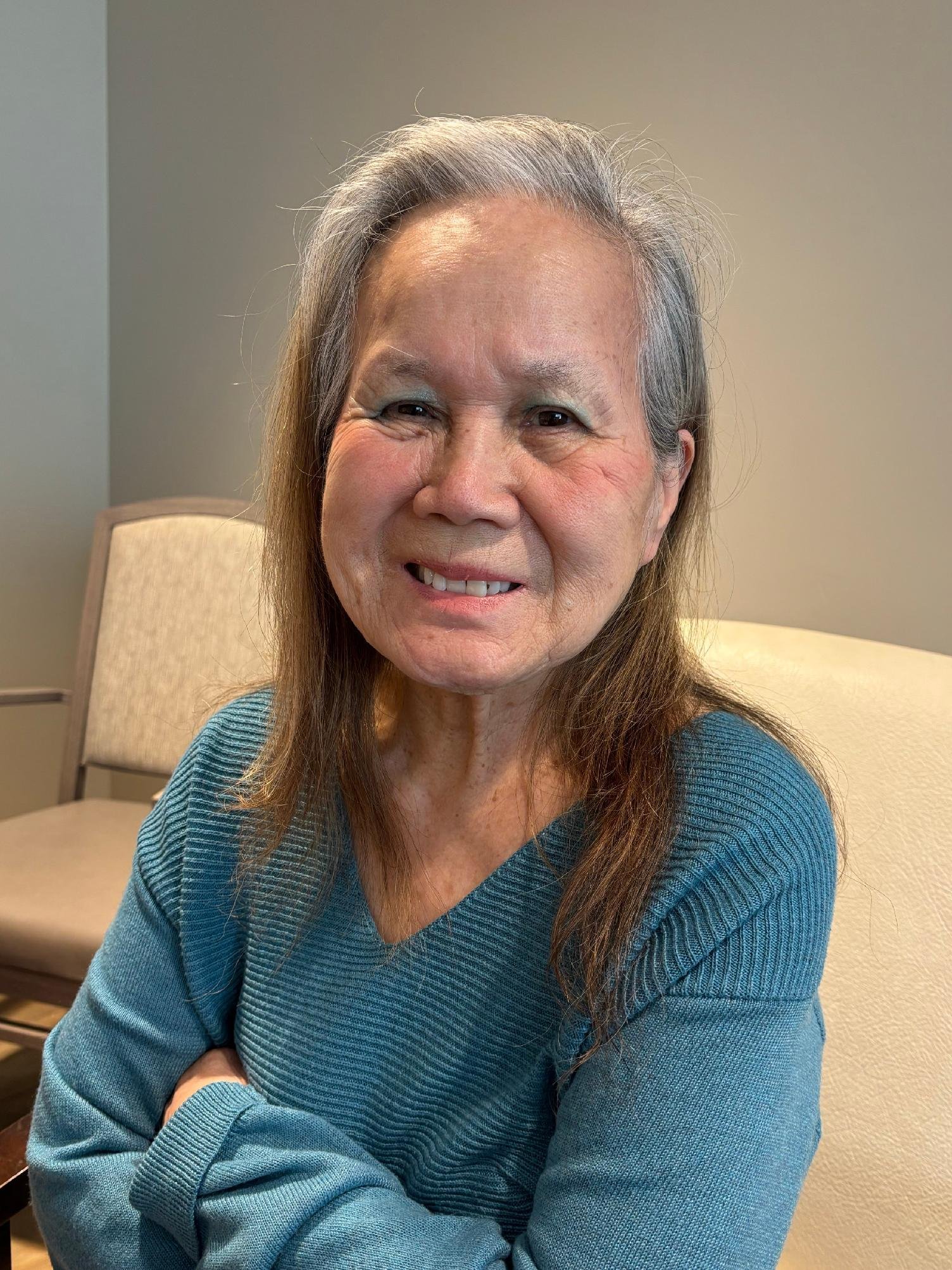 Smiling elderly woman with long gray hair wearing a teal sweater, sitting in a room with beige walls and a chair in the background.