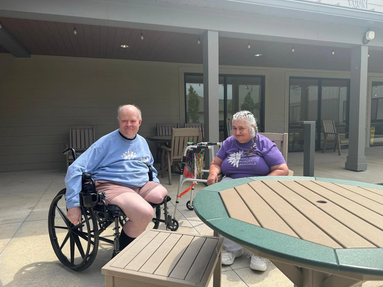 Two elderly people, a man in a wheelchair and a woman with a cane, sitting outdoors at a table on a patio with a building in the background.
