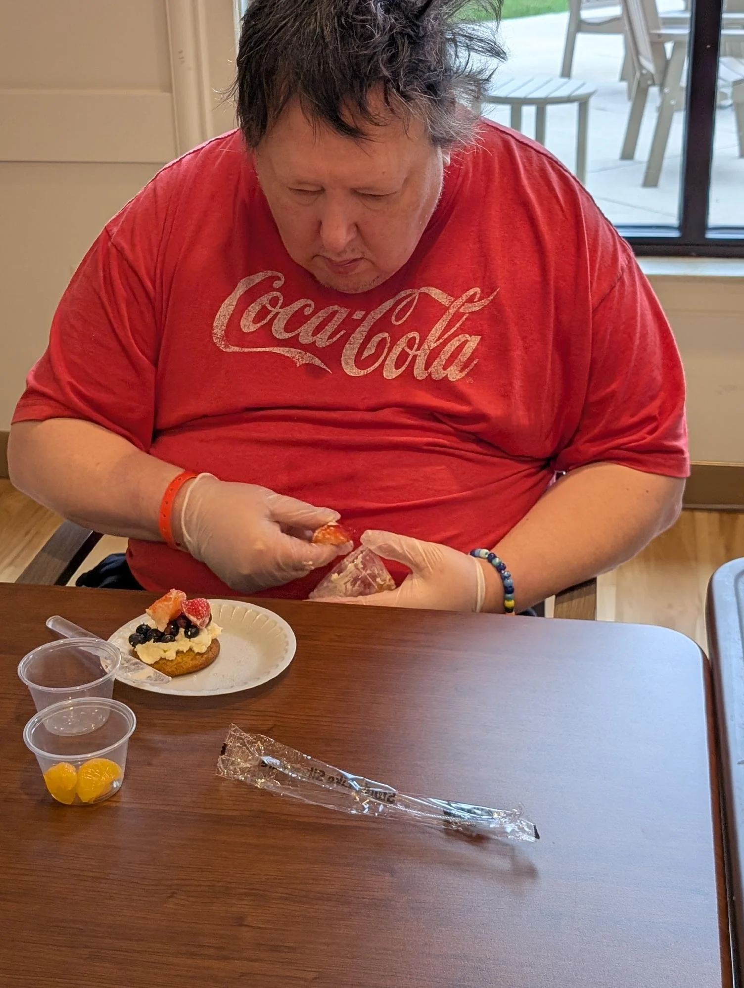 A man wearing a red Coca-Cola T-shirt and latex gloves is preparing or serving a dessert that appears to be a cupcake with frosting and berries. There are small cups of yellow fruit and a plastic-wrapped stir stick on the table in front of him.