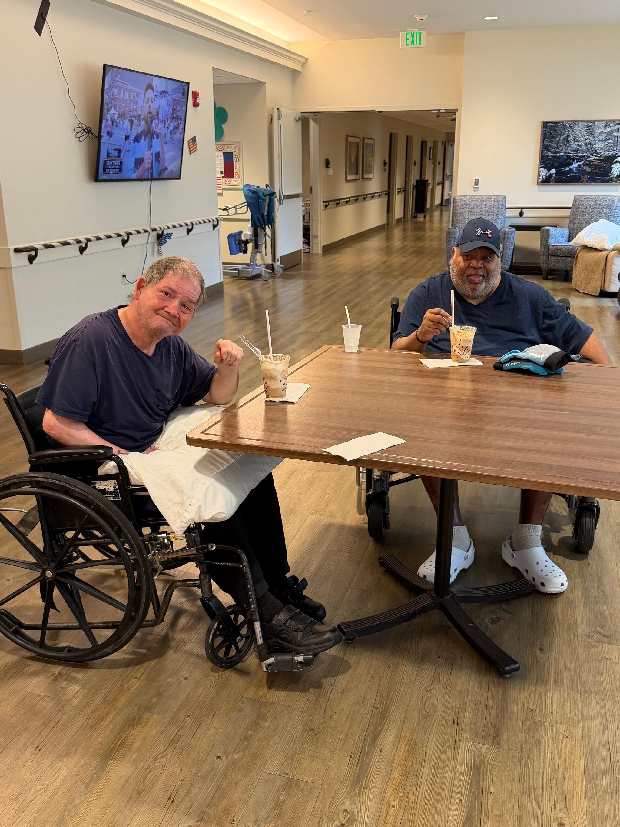 Two men sit at a table in a healthcare facility or nursing home. One man in a wheelchair is making a fist with his right hand, smiling, and the other man is wearing a cap and eating a dessert. The room has wood floors, a TV on the wall, and a hallway