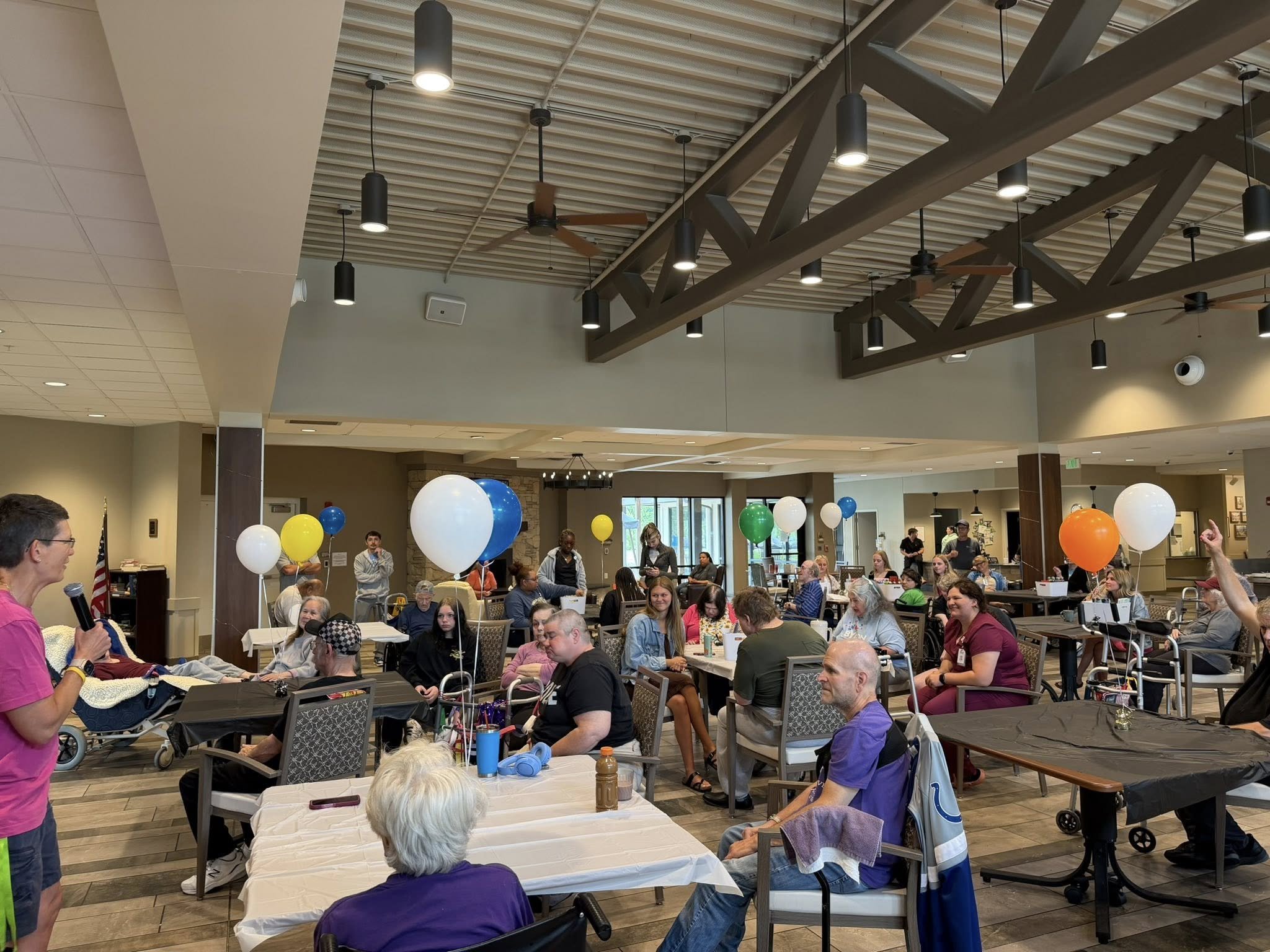 A large indoor gathering at a community event with people seated at tables, balloons for decoration, and a woman speaking at a microphone, with others interacting and walking around.