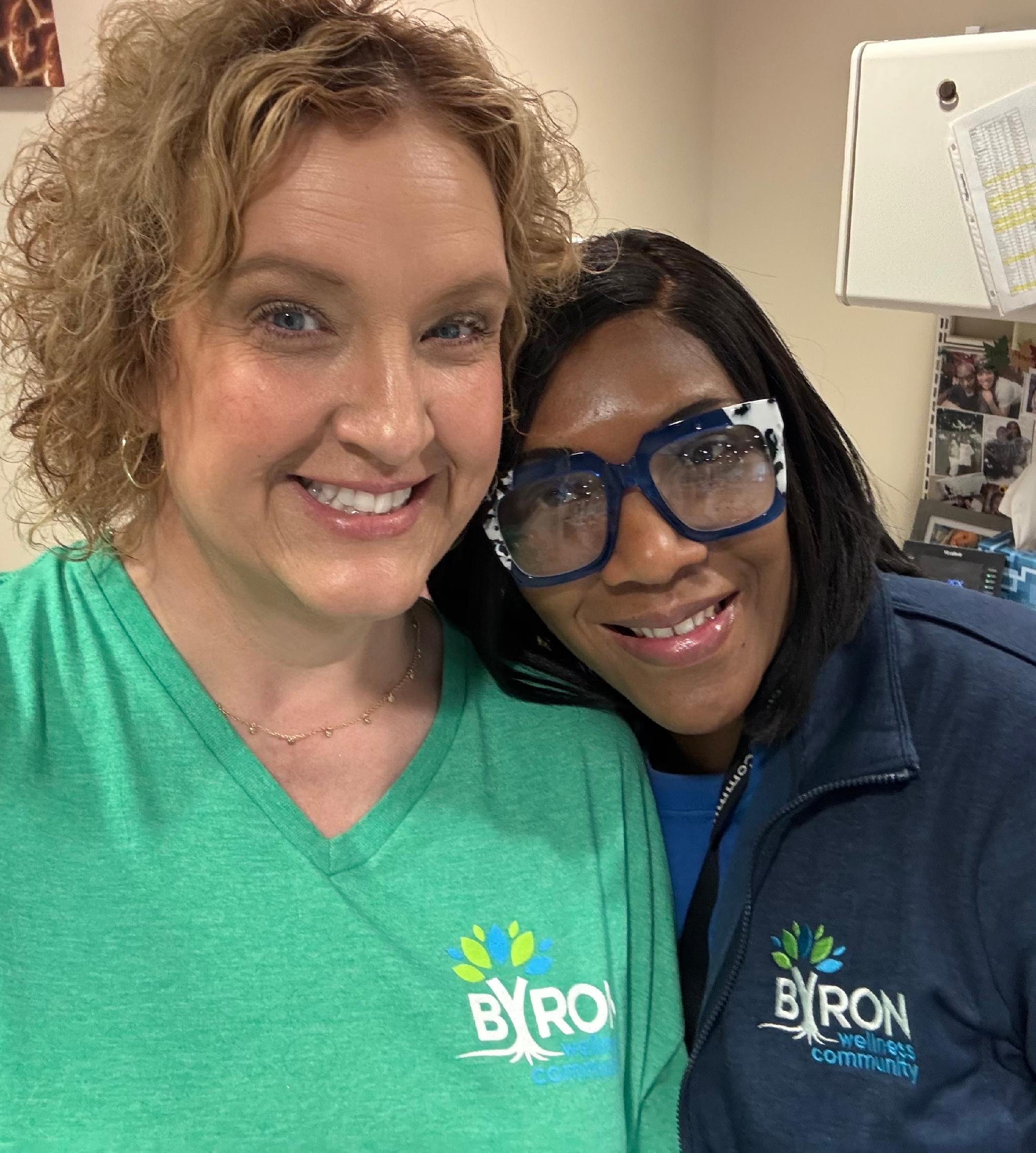 Close-up of two women smiling, standing together indoors, wearing BYRON community shirts with a tree and text on them, in front of a wall with photos and papers pinned up.