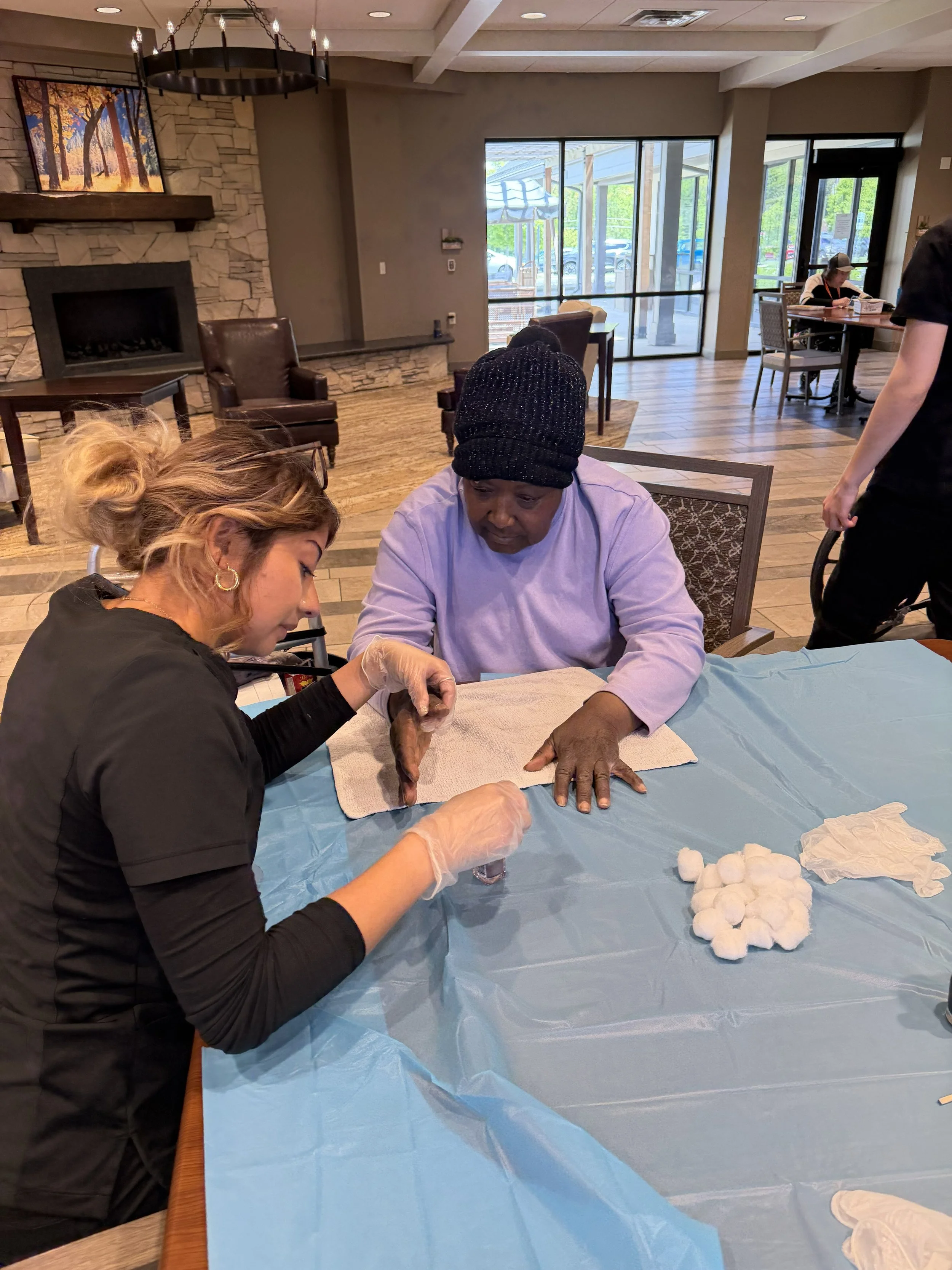 Two women are sitting at a table, one of them is receiving a tattoo on her hand while the other assists. The setting appears to be a community center or a similar indoor space, with wooden floors, chairs, a stone fireplace, and large windows showing 