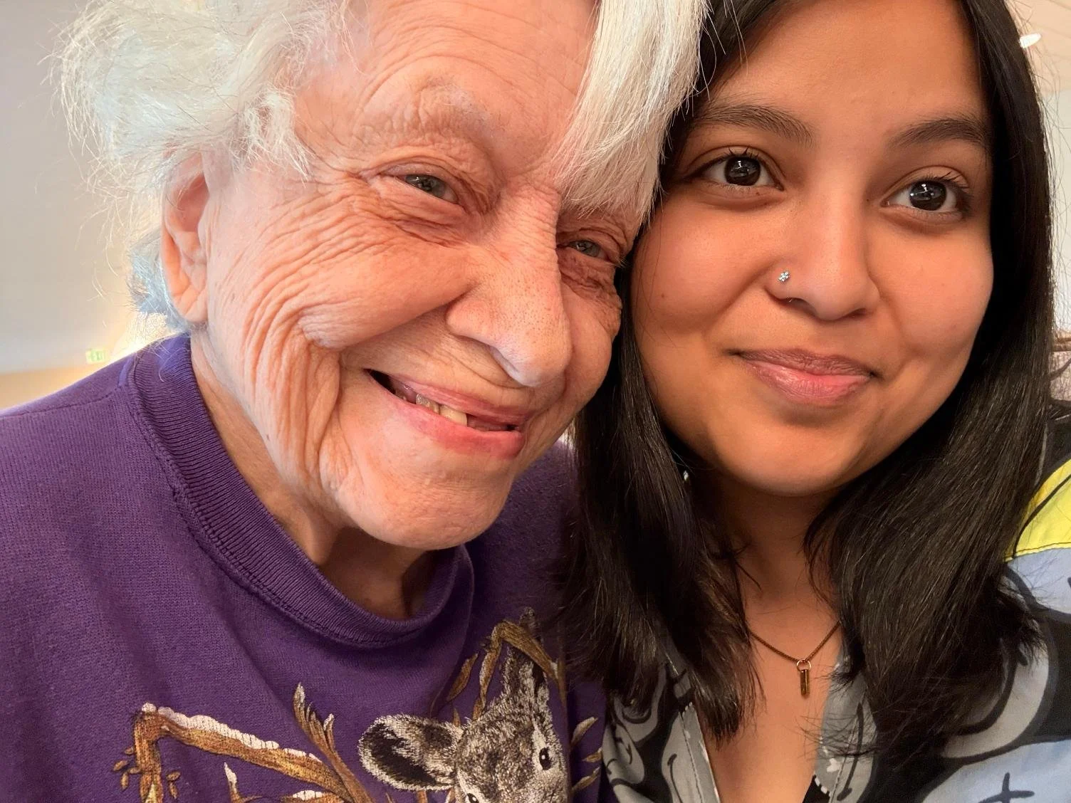Close-up of an elderly man with white hair smiling with a young woman with dark hair, both posing closely for a selfie indoors.