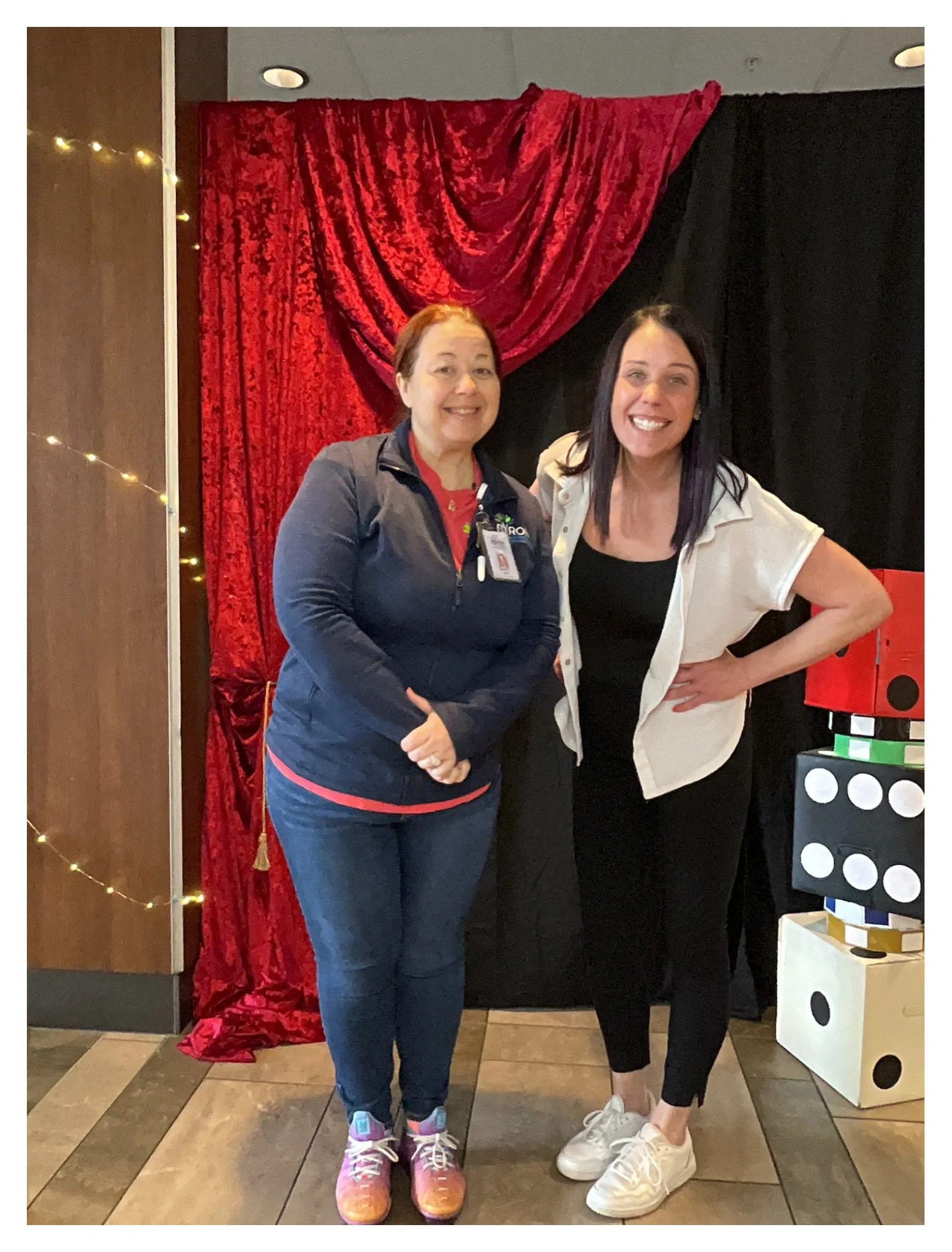 Two women standing together in a restaurant near a decorative red and black curtain backdrop. One woman wears a navy jacket and colorful sneakers, and the other wears a white shirt over a black top with white sneakers. They are smiling and posing for