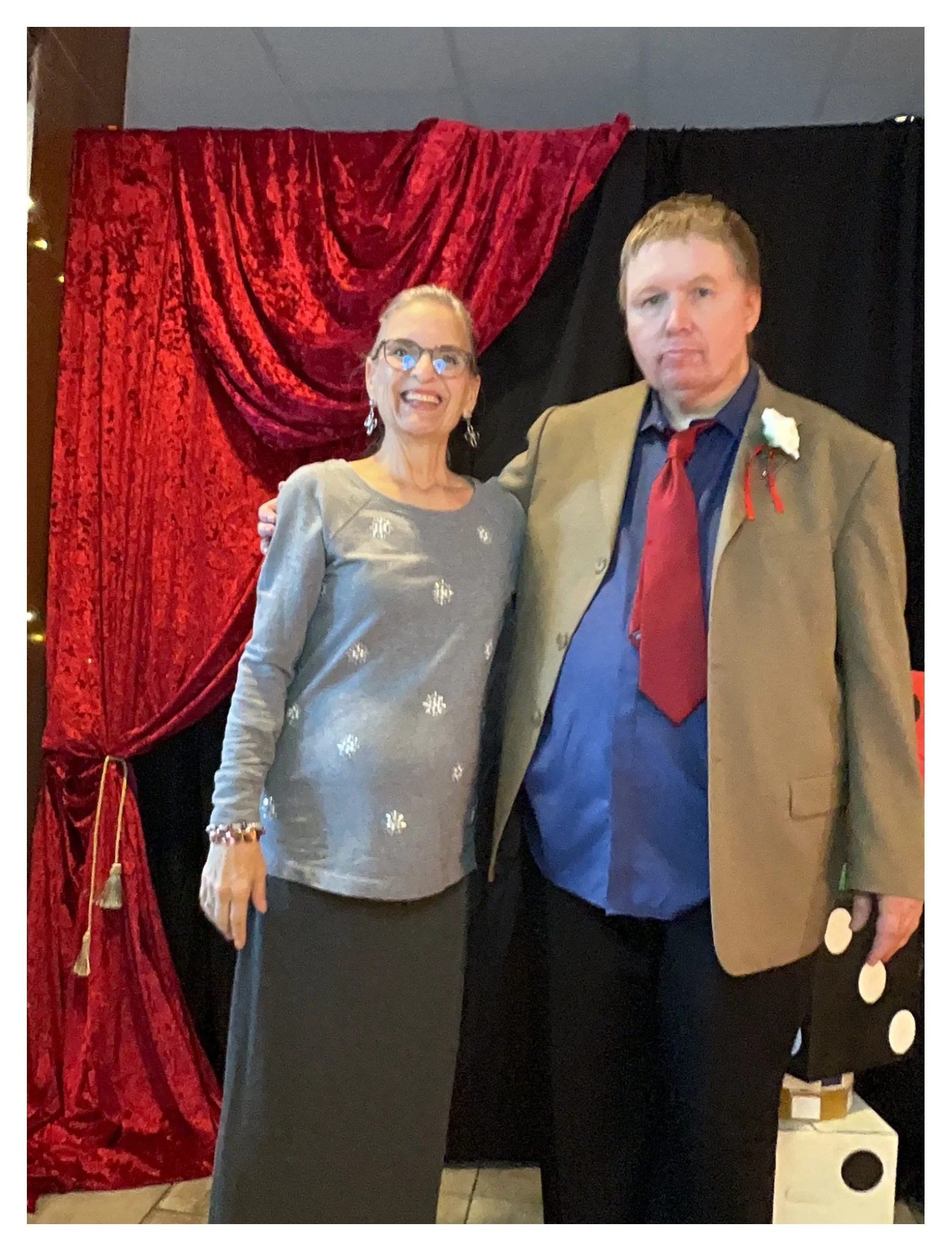 Two people standing in front of red and black draped fabric backdrop, smiling, with formal attire and corsages, at what appears to be a celebration or event.