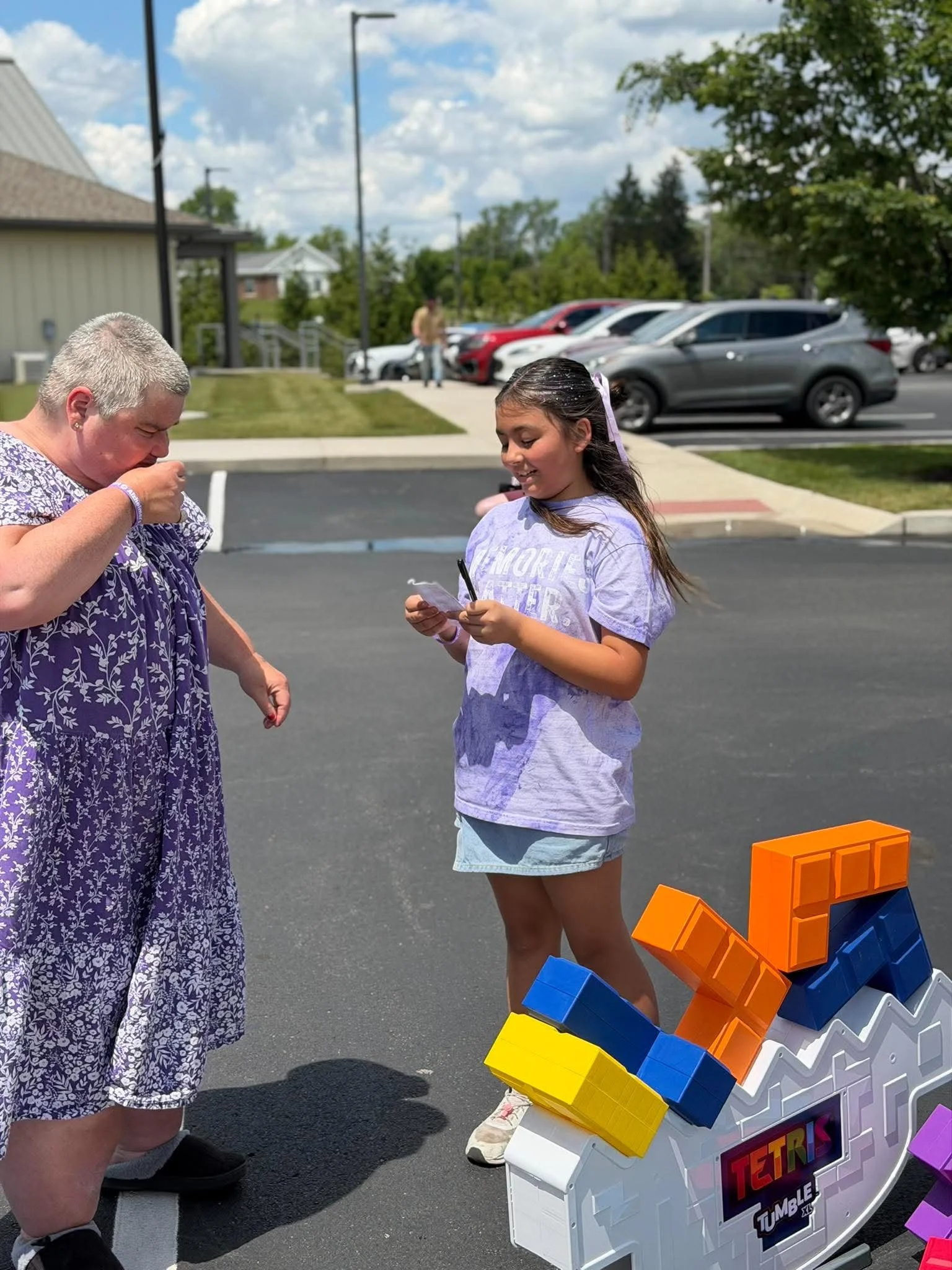 Two young girls with dark hair and light skin are playing Tetris Tumble game outside on a sunny day. One girl has long hair with a purple ribbon and is holding a phone, smiling, and the other girl has short hair with platinum blonde and is wearing a 
