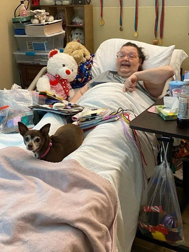 A woman lying in a hospital bed smiling, surrounded by medical equipment and stuffed animals; a small dog sitting on the bed near her, with a pink collar and looking at the camera.