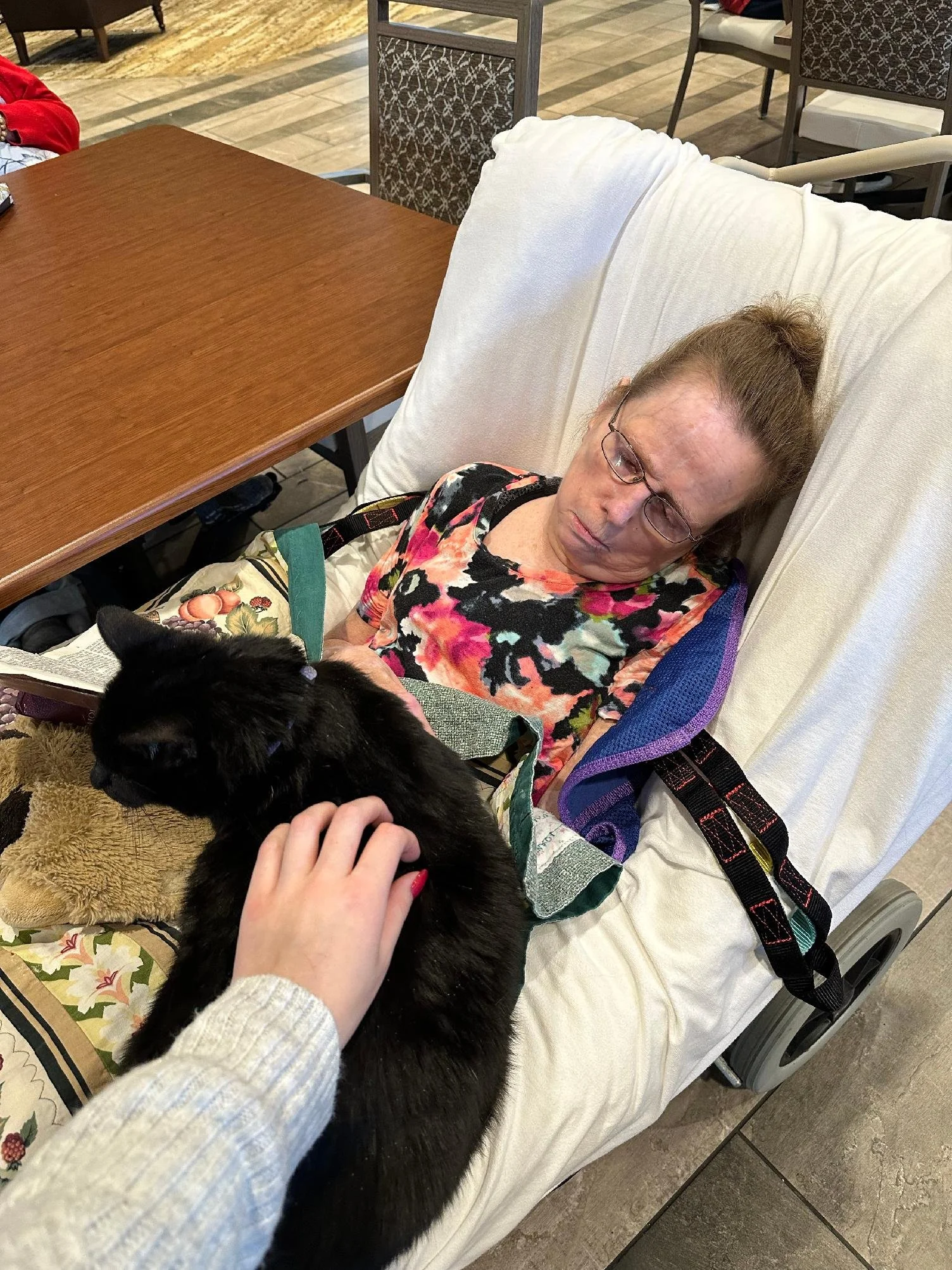 An elderly woman with glasses and a bun hairstyle sleeping in a hospital bed, with a black dog lying next to her and a person petting the dog. The bed has a white pillow and a blanket, and the woman is wearing a colorful floral shirt.