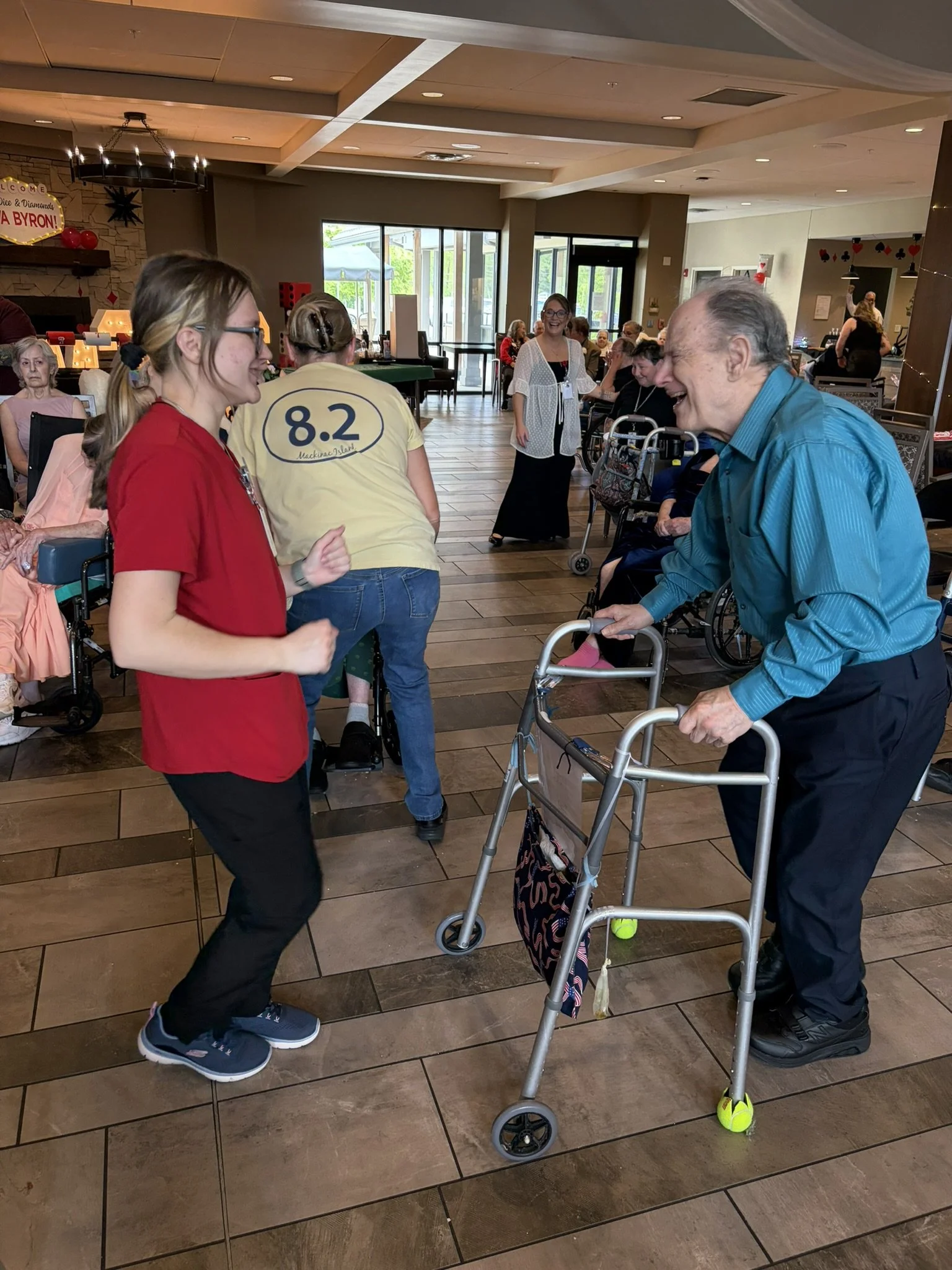 A group of people, including elderly individuals and younger women, are gathered in a bright, spacious room, engaging in a lively social activity. Several participants have mobility aids like walkers, and the atmosphere appears joyful and friendly.