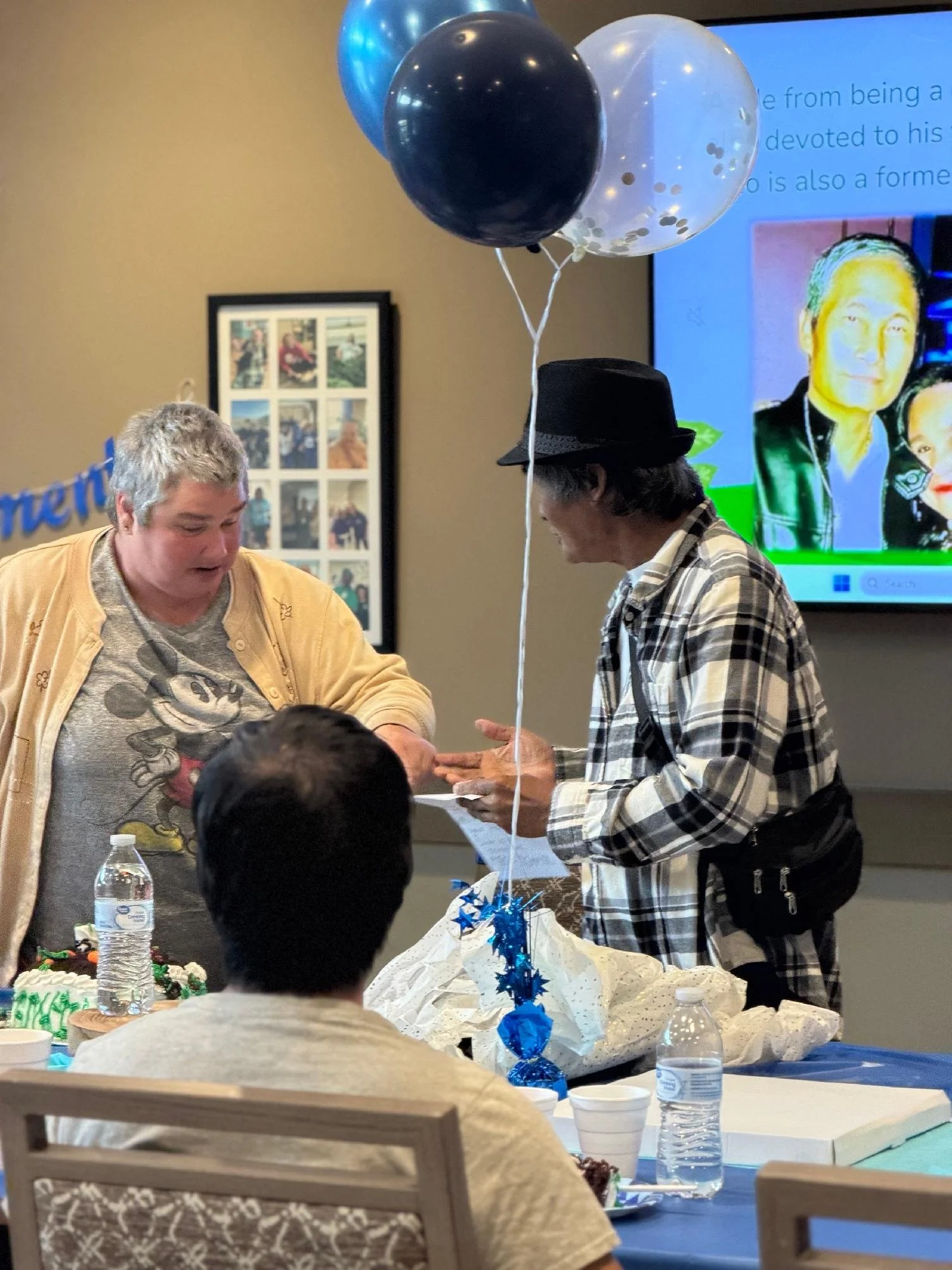 A birthday party scene with a decorated table, balloons, and people. One person is helping another with a gift or card. There is a cake on the table and a celebration.