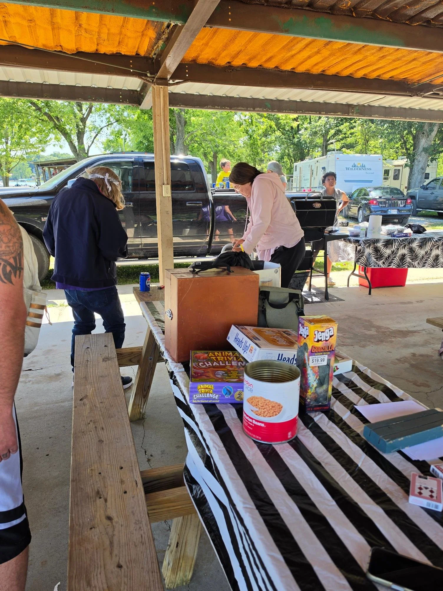 People gathered at a barbecue or picnic area with tables and food, including canned beans, board games, and a grill, under a covered shelter with vehicles and trees in the background.