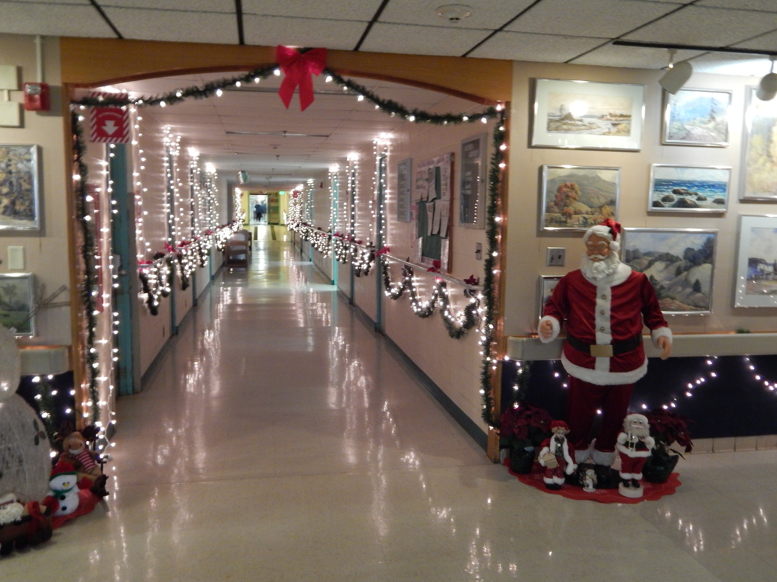 A hallway decorated for Christmas with string lights, wreaths, and ribbons. A Santa Claus figure and smaller holiday decorations are placed on the right side.