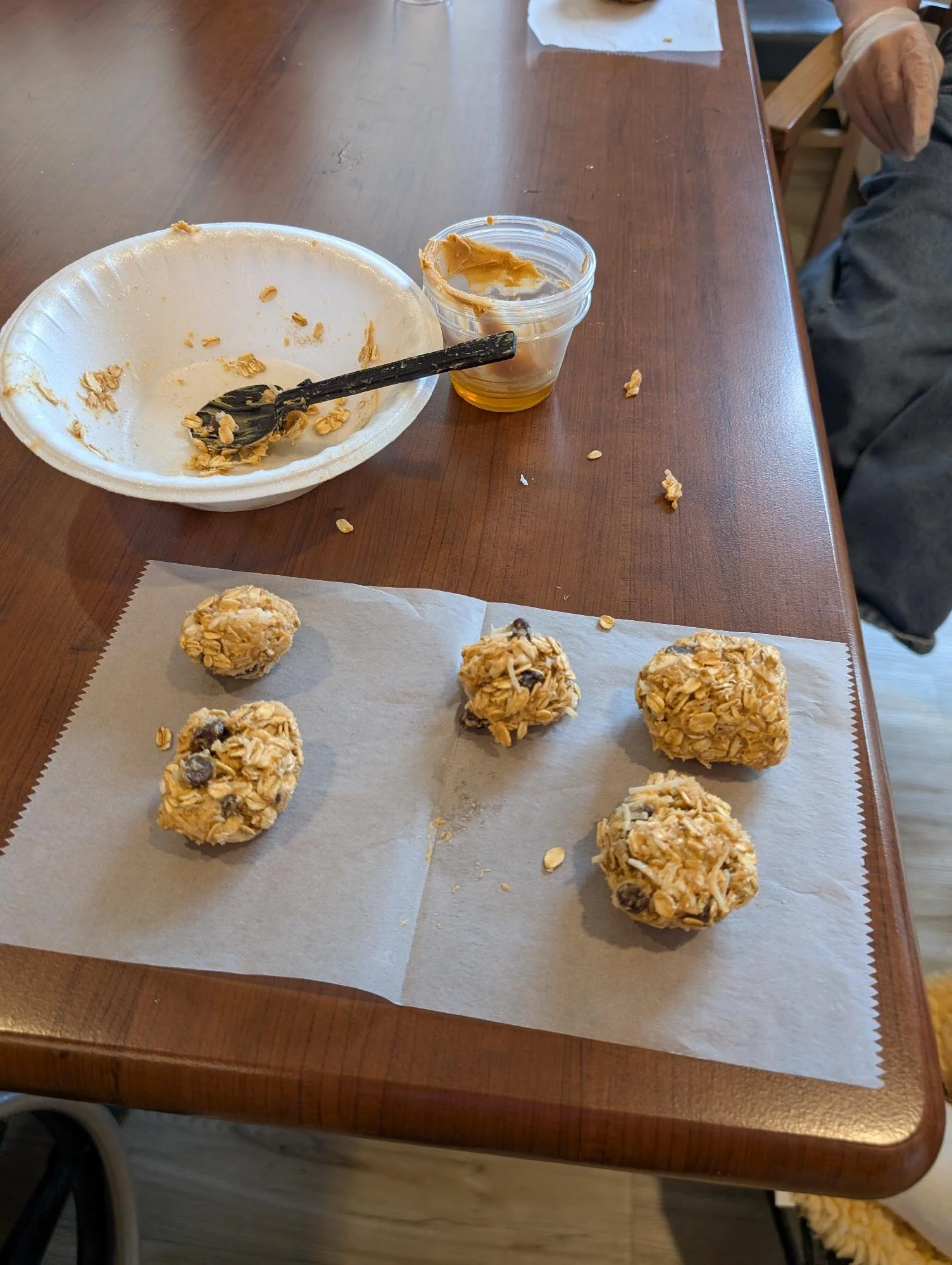 Oatmeal cookie balls on a parchment-lined tray, with crumbs and a bowl of remaining mixture nearby on a wooden table.