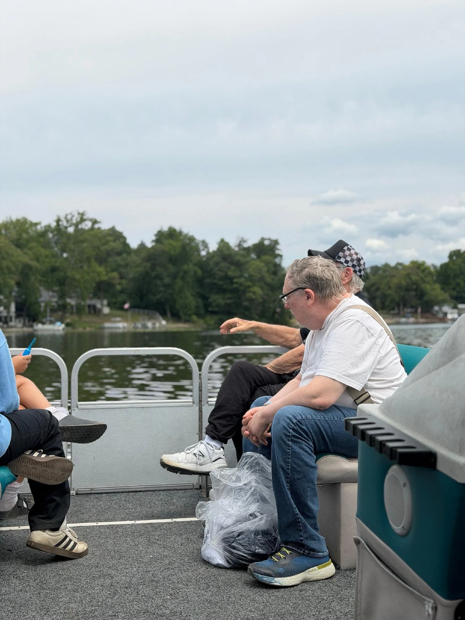 People sitting on benches outdoors near a body of water, with trees in the background, under a cloudy sky.