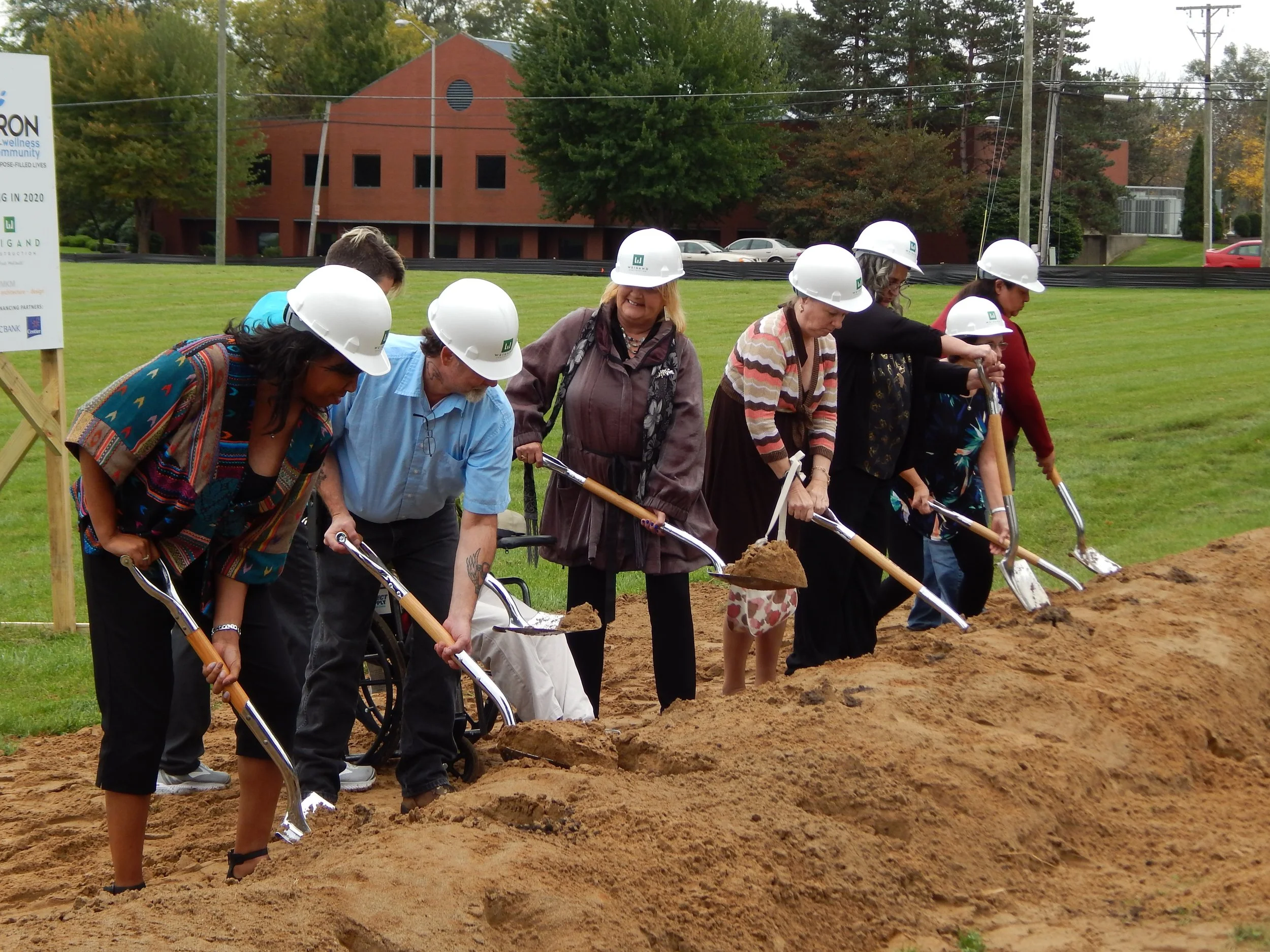 Group of people wearing white hard hats and holding shovels, participating in a groundbreaking ceremony on a grassy field with a building and trees in the background.