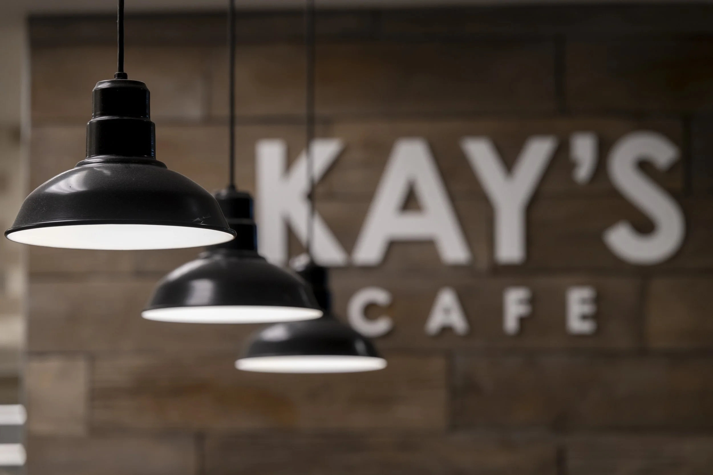 Multiple black hanging pendant lights in a row inside a cafe with a wooden wall in the background, which has large white letters spelling out parts of the word 'KAYS'.