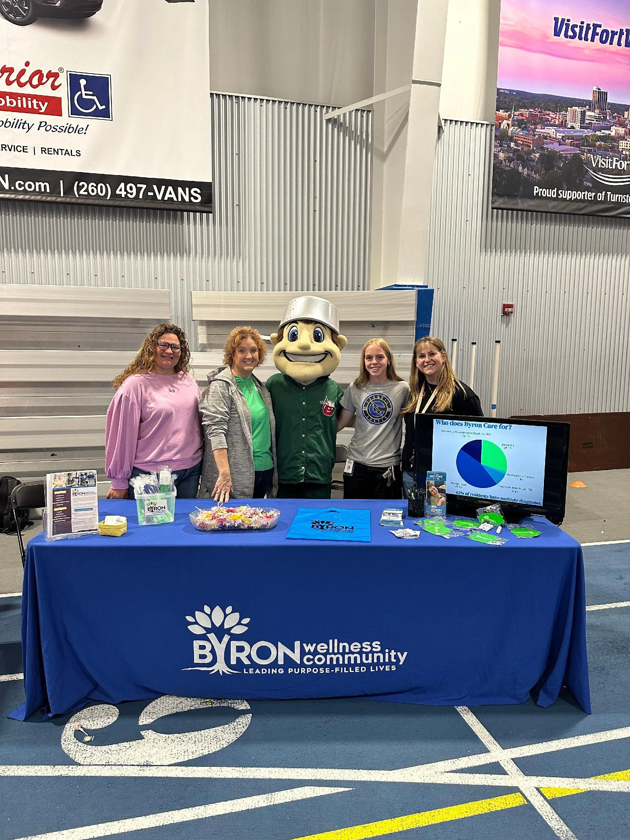 Table with a blue tablecloth displaying the BYRON wellness community logo, brochures, and promotional items, set up in a parking lot with white and yellow parking lines visible on the ground.