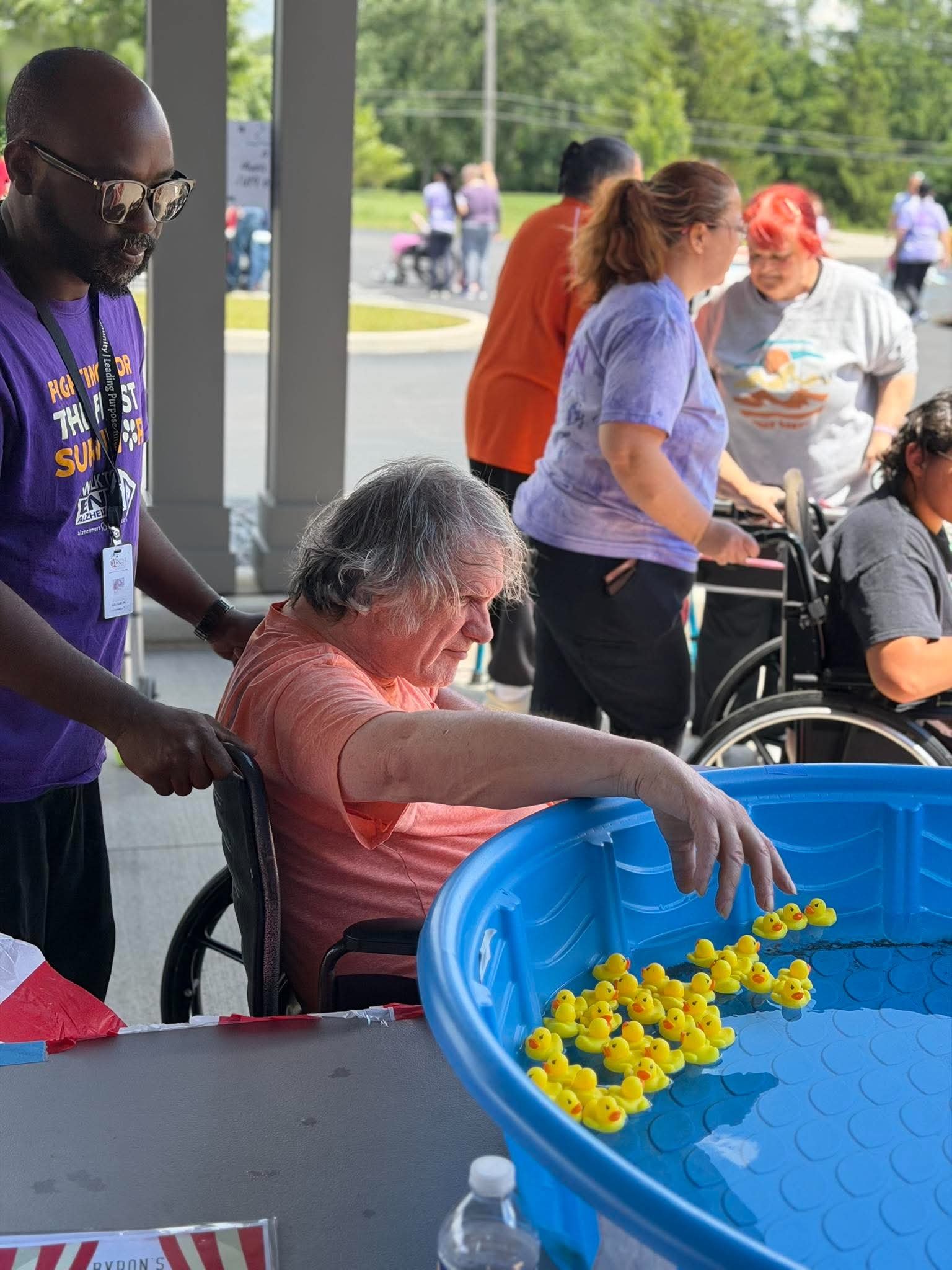 A person in a wheelchair reaches into a large blue tub filled with yellow rubber ducks at an outdoor event, with several people and green trees in the background.