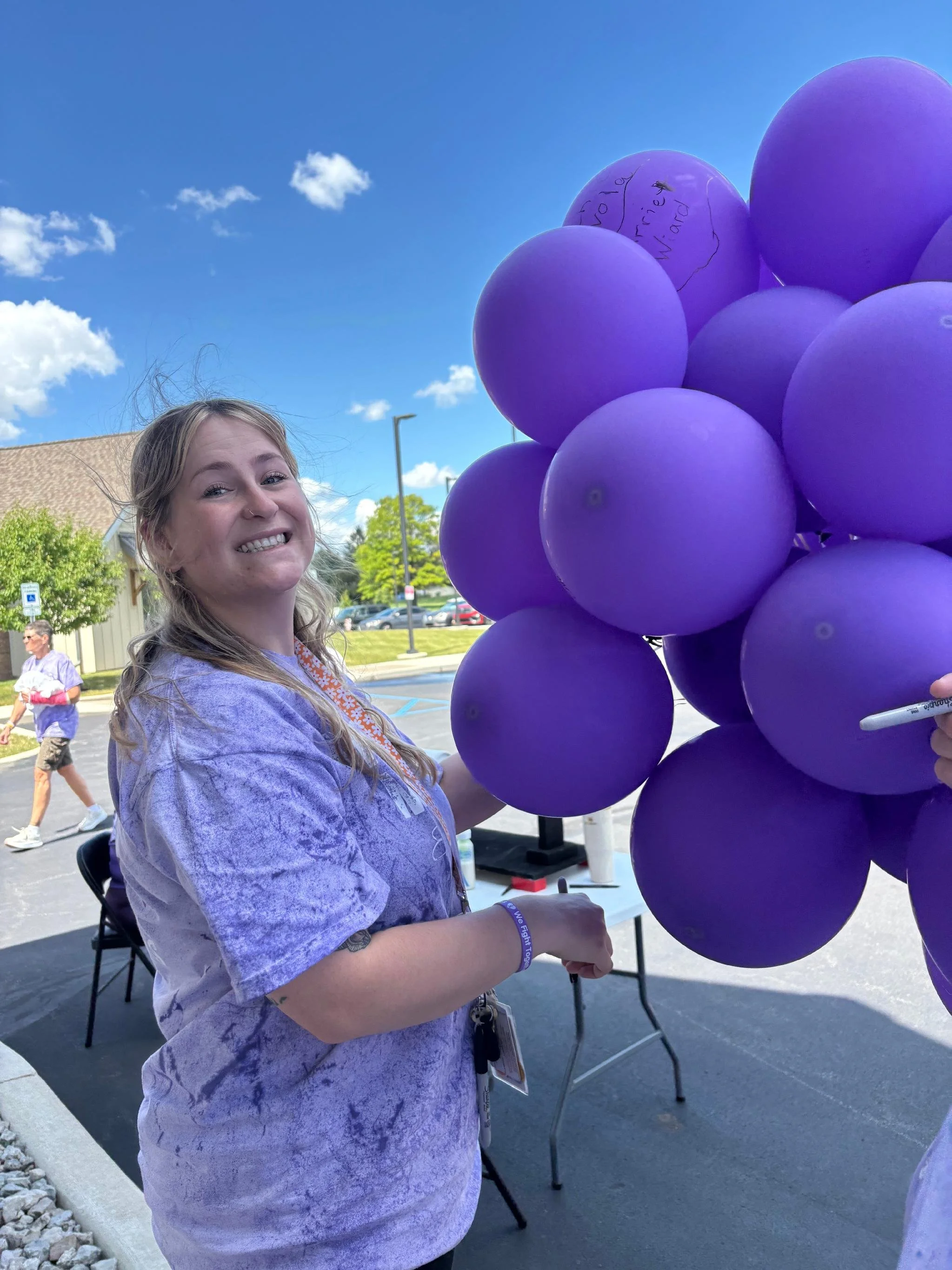 A young woman with long hair and a purple tie-dye shirt smiling while holding a bunch of purple balloons outside on a sunny day. There are other people visible in the background and a building with a parking lot.