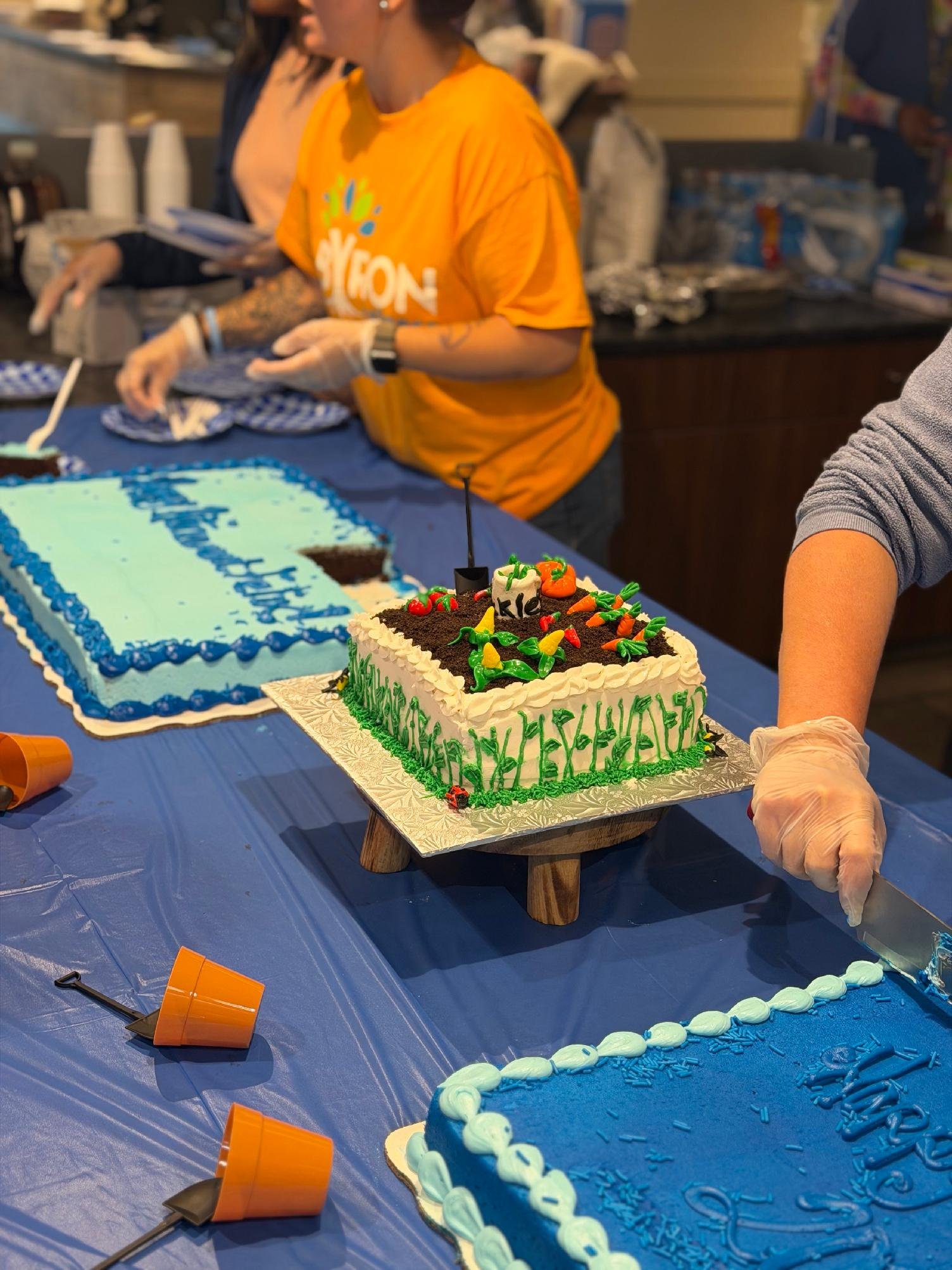 A cake decorated with green and yellow icing, colorful candies, and a small candle, on a wooden stand at a celebration. A woman in an orange shirt is slicing another cake on a table covered with a blue tablecloth, surrounded by other people celebrati