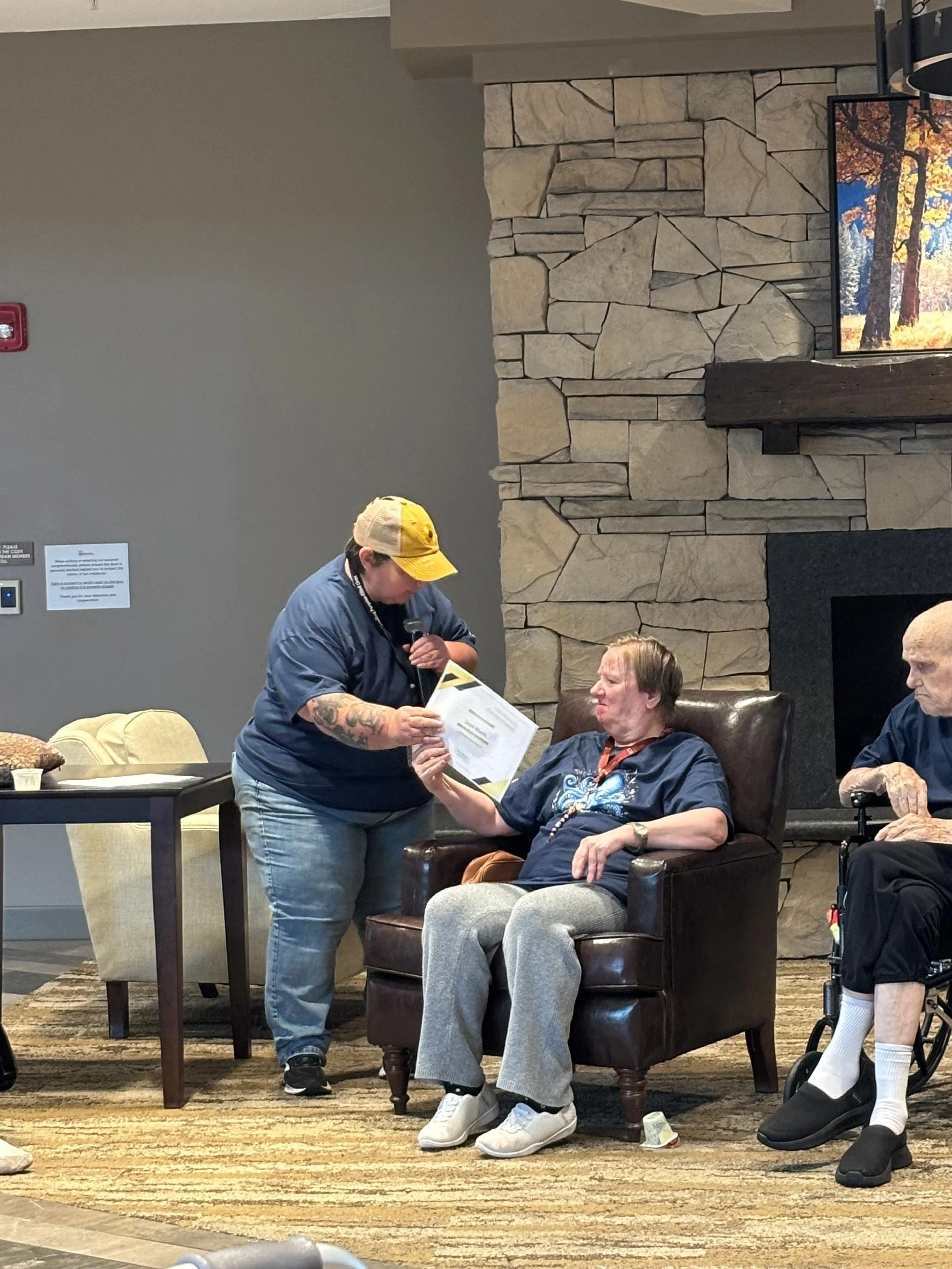 A woman sitting in an armchair receives a hand massage from a man standing beside her. There are elderly people in the background, a stone fireplace, a mounted TV, and a fireplace mantle. The setting appears to be a community or healthcare facility.