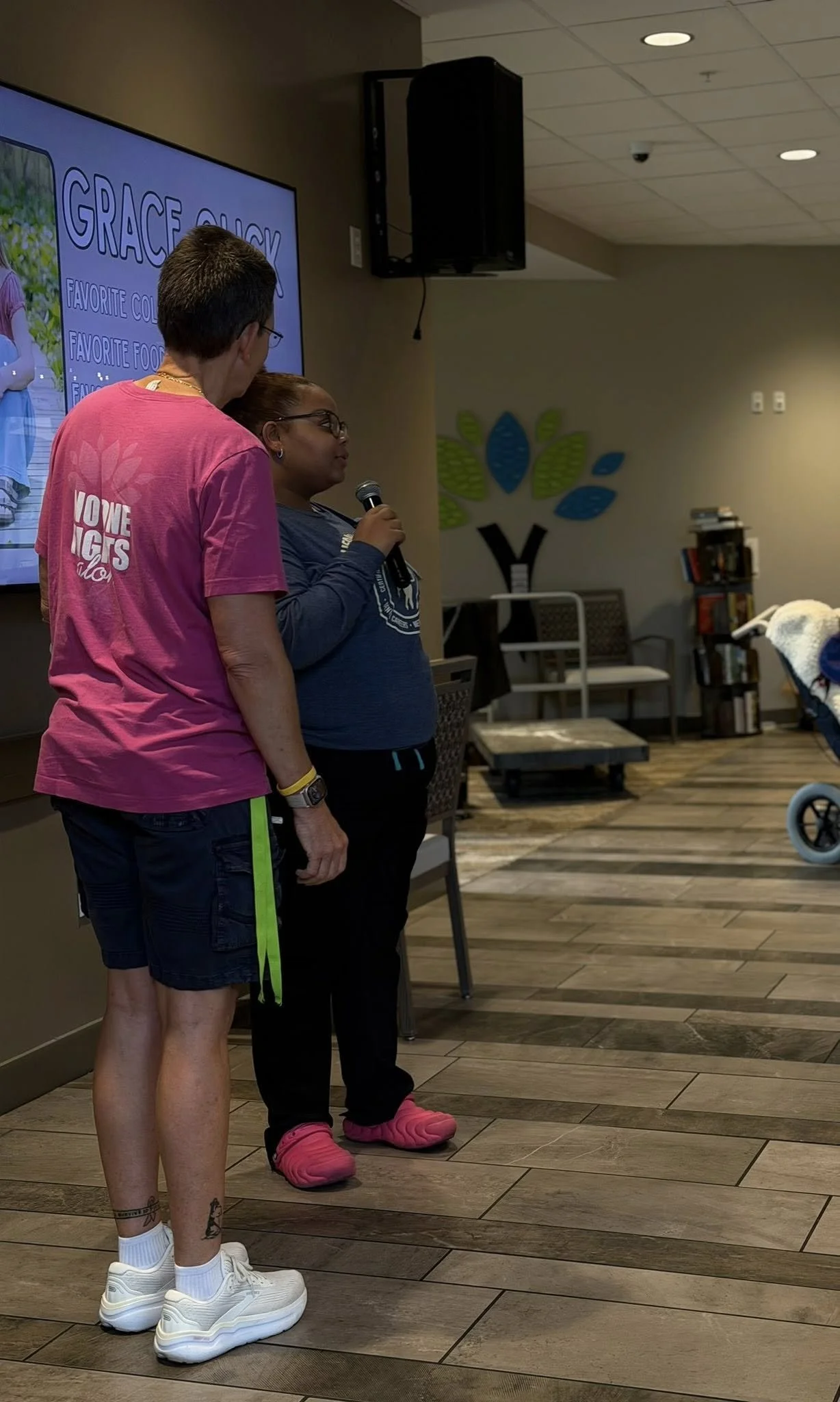 A woman and a girl are standing indoors near a large TV screen. The woman is wearing a pink T-shirt, shorts, white sneakers, and has tattoos on her legs. The girl is wearing glasses, a blue long-sleeve shirt, black pants, and pink slippers, and is ho
