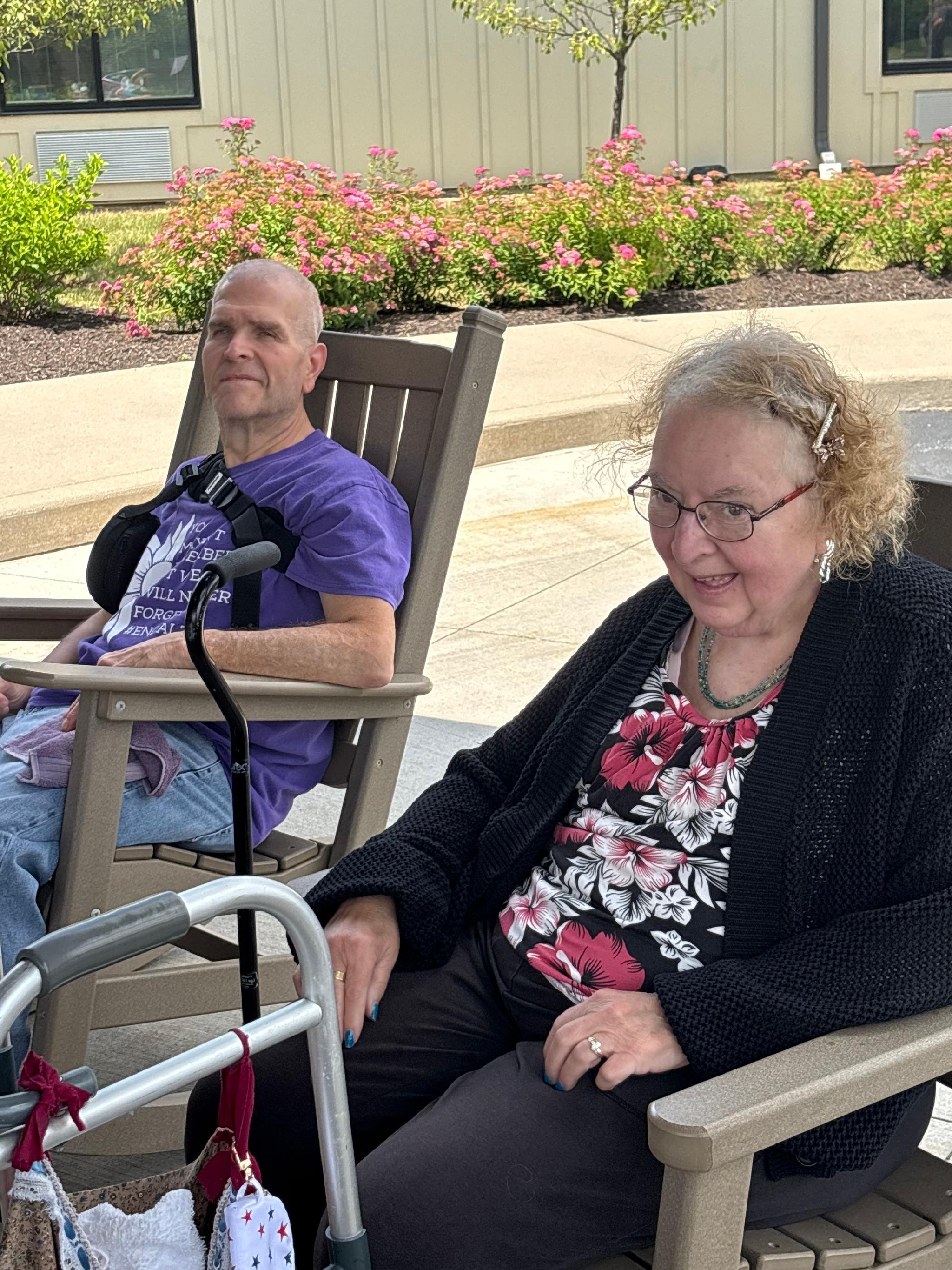A man in a purple shirt sitting in a chair outdoors, a woman in a floral shirt and black jacket sitting beside him, with a walker and pink flowers in the background.