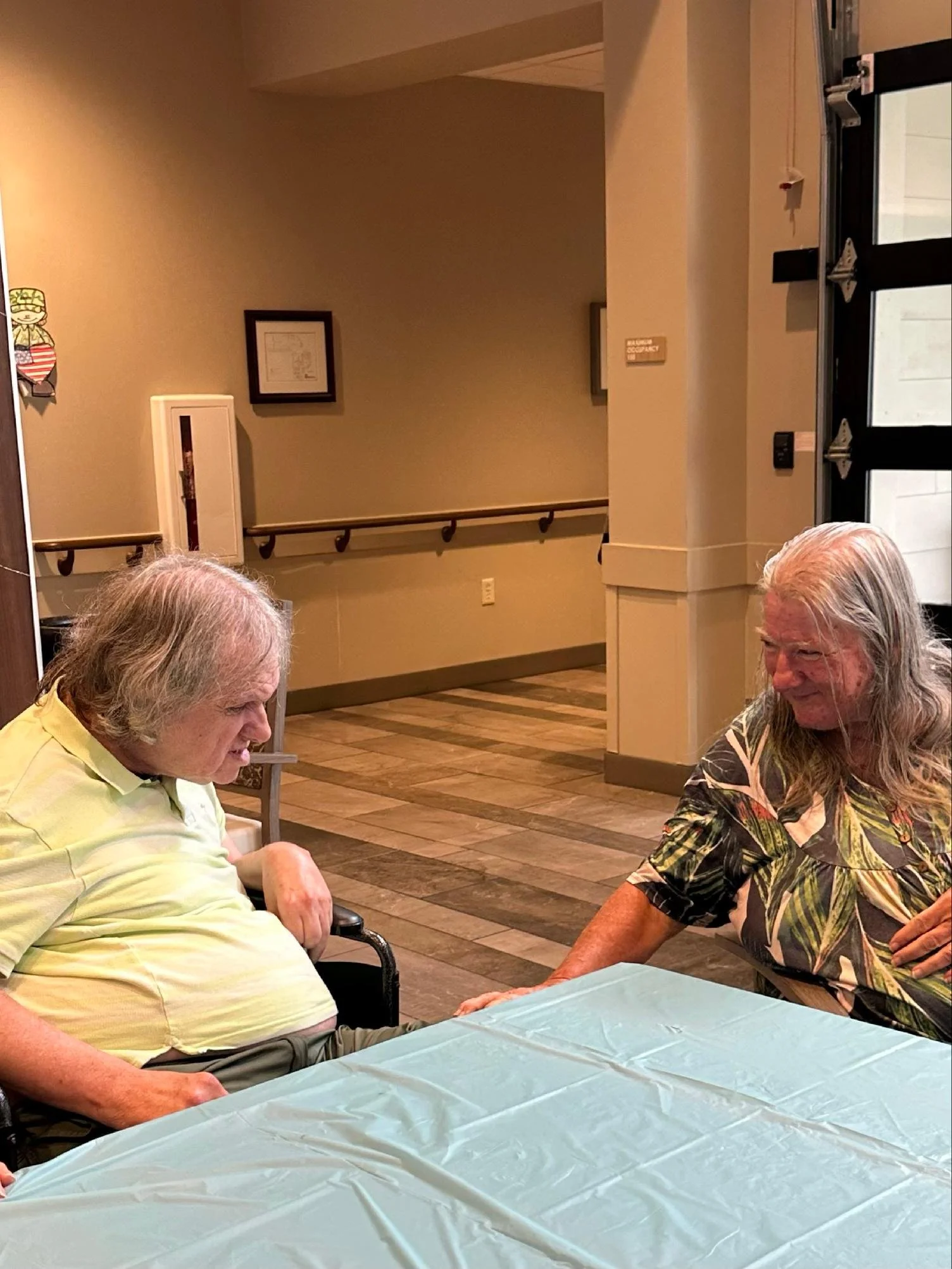 Two elderly men sitting at a table covered with a light blue tablecloth, one wearing a yellow shirt and the other with long gray hair and a patterned shirt, engaged in conversation inside a warmly lit room with wooden floors and framed pictures on th