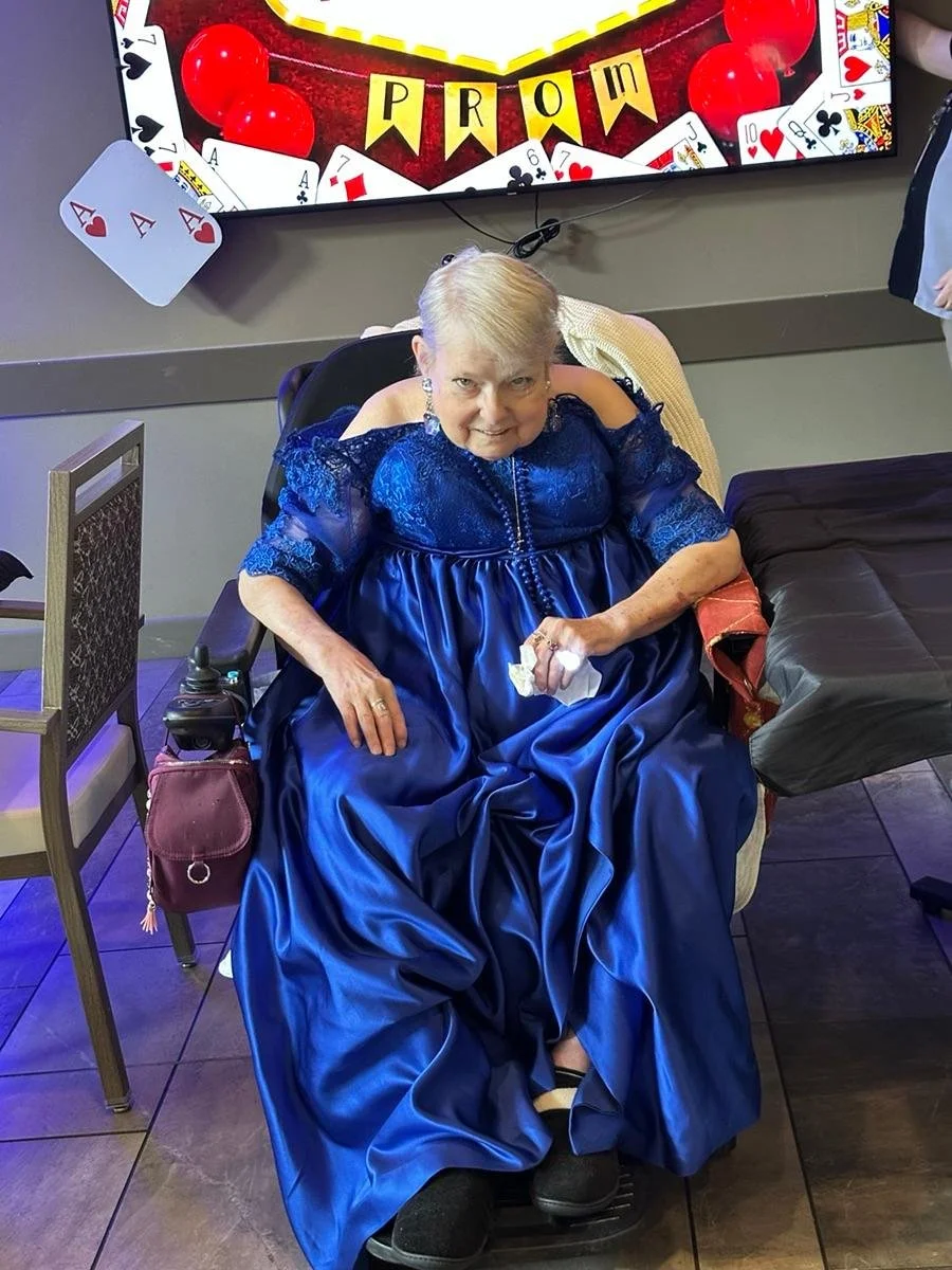 An elderly woman with short blonde hair dressed in a bright blue satin gown sitting in a chair. She is smiling and holding a tissue in her hand. Behind her is a screen displaying a casino-themed message with playing cards and chips. There is a large 