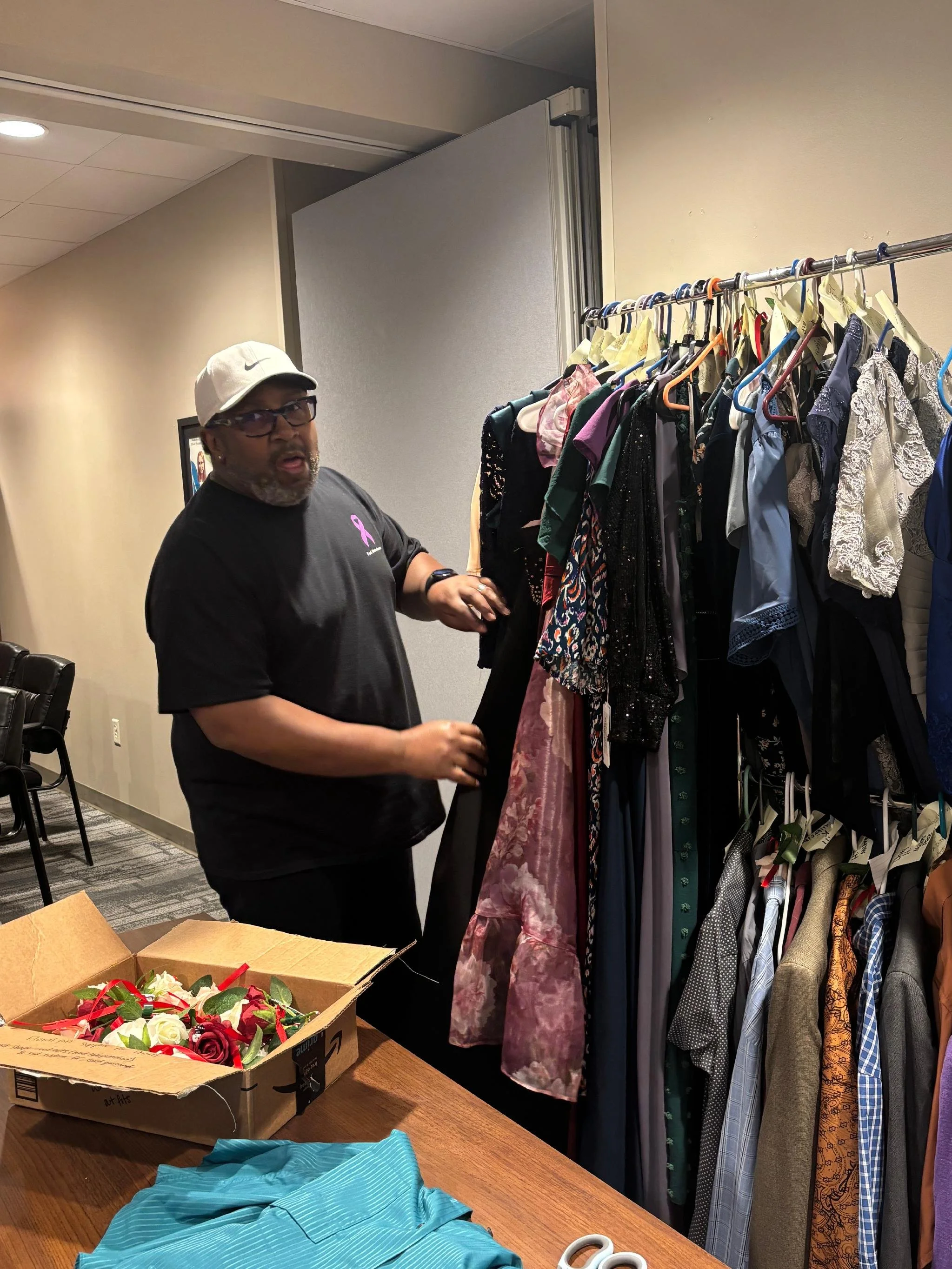 A man wearing glasses, a gray cap, and a black T-shirt with a pink ribbon stands next to a hanging rack of various clothes, possibly at a charity or thrift store. On the table in front of him is an open box of artificial flowers and some blue fabric,