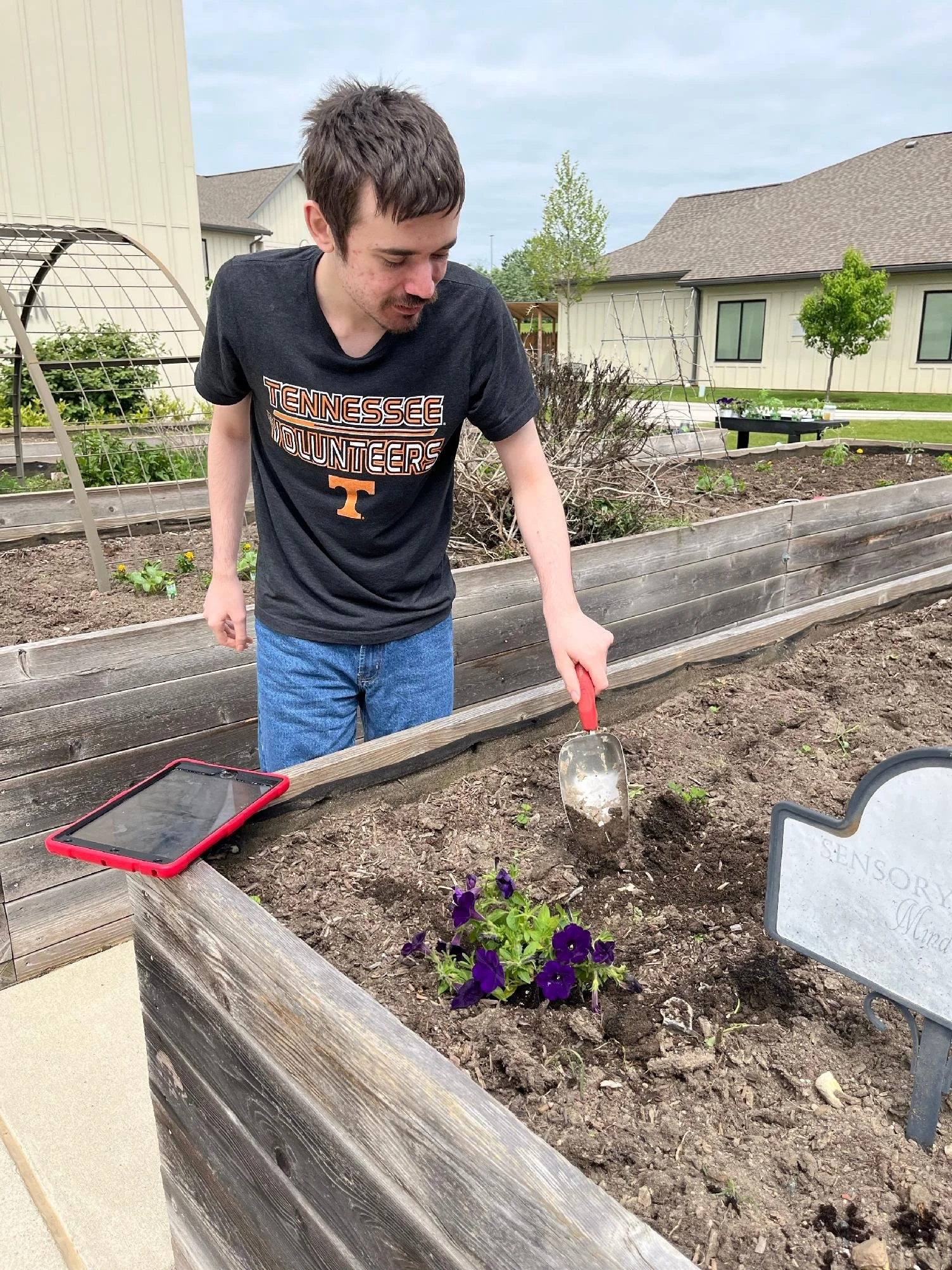 A young man with dark hair and a goatee gardening in a raised garden bed, holding a small gardening trowel, in a suburban backyard with houses and trees in the background.