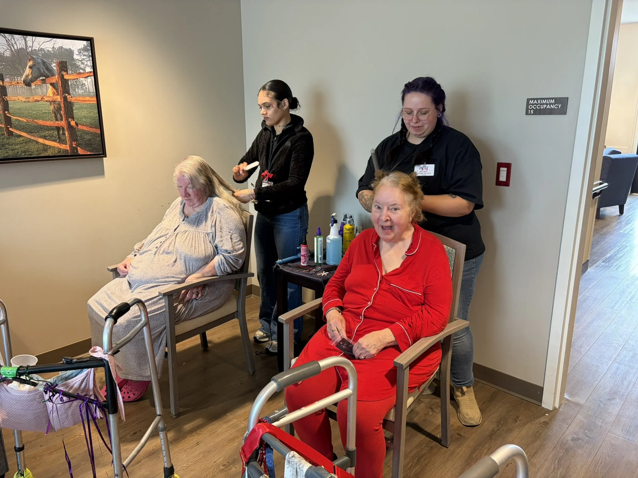 Two elderly women sitting in chairs, receiving haircuts from two women standing behind them. The room has beige walls, with a framed painting of a horse on a fence on the left. A table next to the women has hair styling supplies.