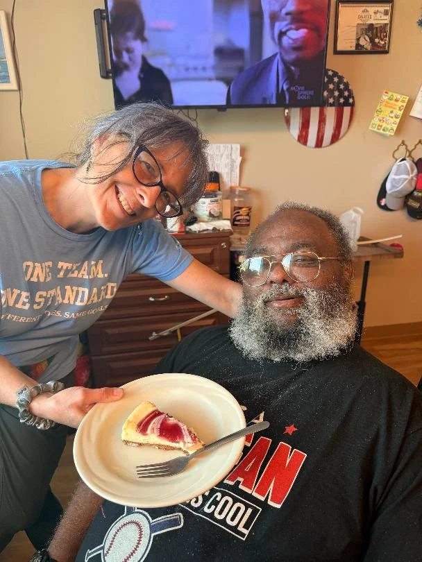 A woman with glasses and a blue T-shirt smiling and leaning over an elderly man with glasses and a gray beard, who is sitting while holding a plate with a slice of cherry cheesecake. They are indoors with a television playing in the background.