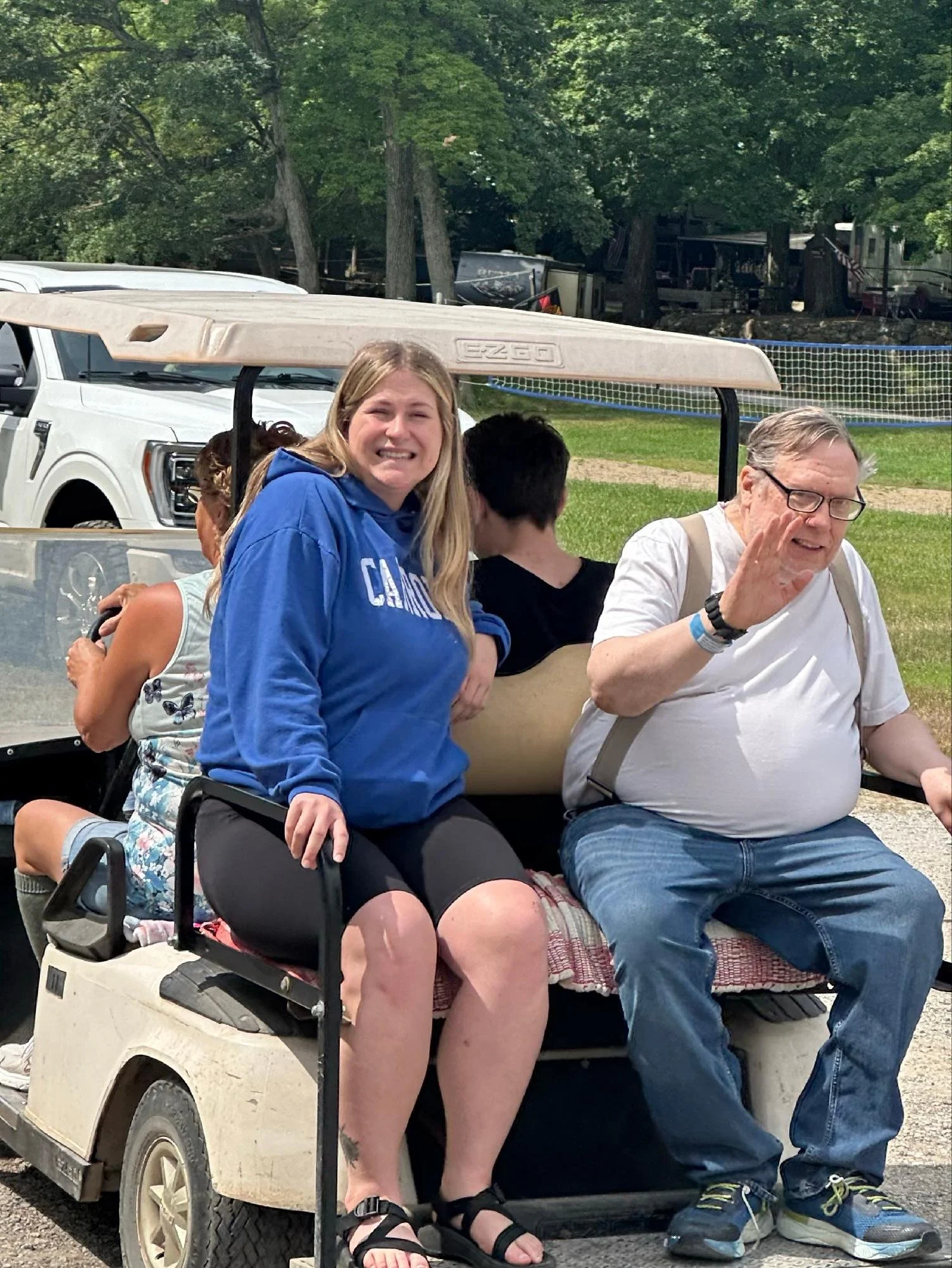 People sitting on a golf cart outdoors, with a woman in a blue hoodie smiling, an older man waving, and other individuals in the background, surrounded by trees and green grass.