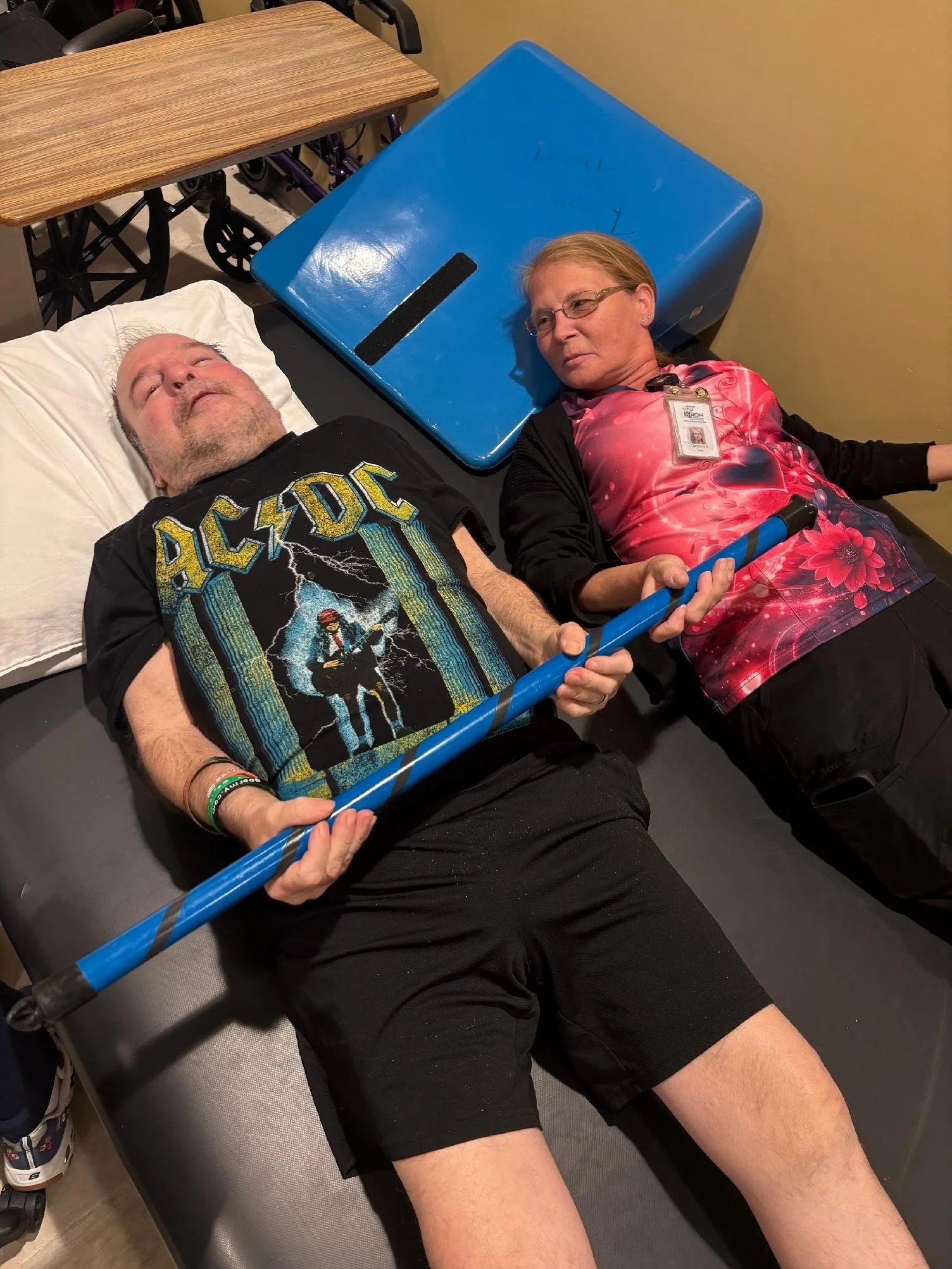 A man lying on a hospital bed with an oxygen mask on, holding a blue walking stick, and a woman sitting beside him on the bed, resting her head on a blue table, and holding onto the walking stick.
